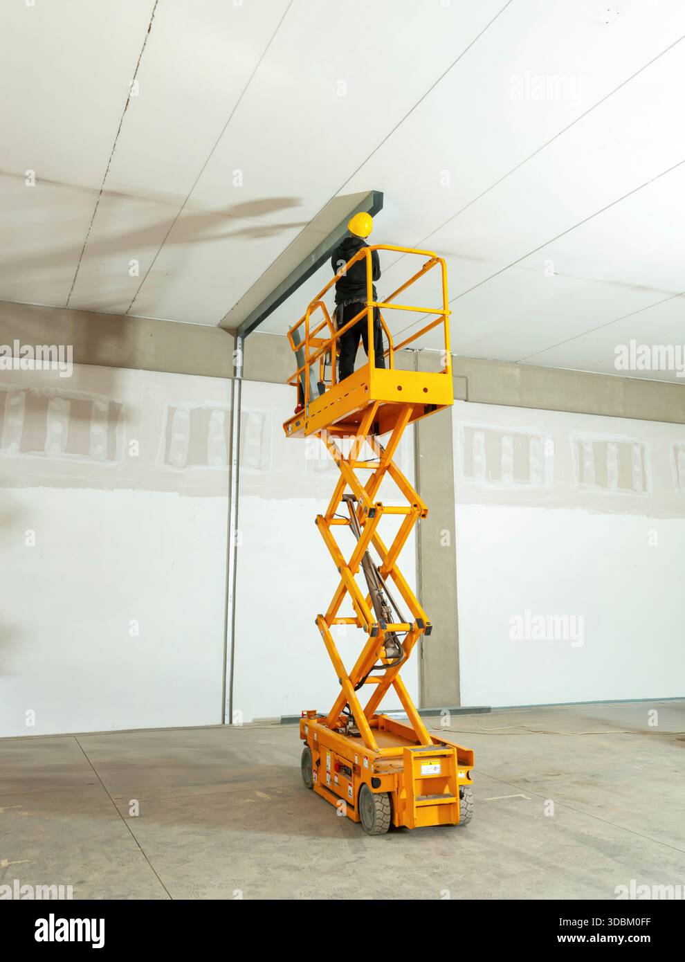 Il lavoratore edile indossa un casco di sicurezza che utilizza una piattaforma di lavoro mobile elevabile per installare travi a soffitto in un magazzino durante lavori di ristrutturazione o lavori edili Foto Stock