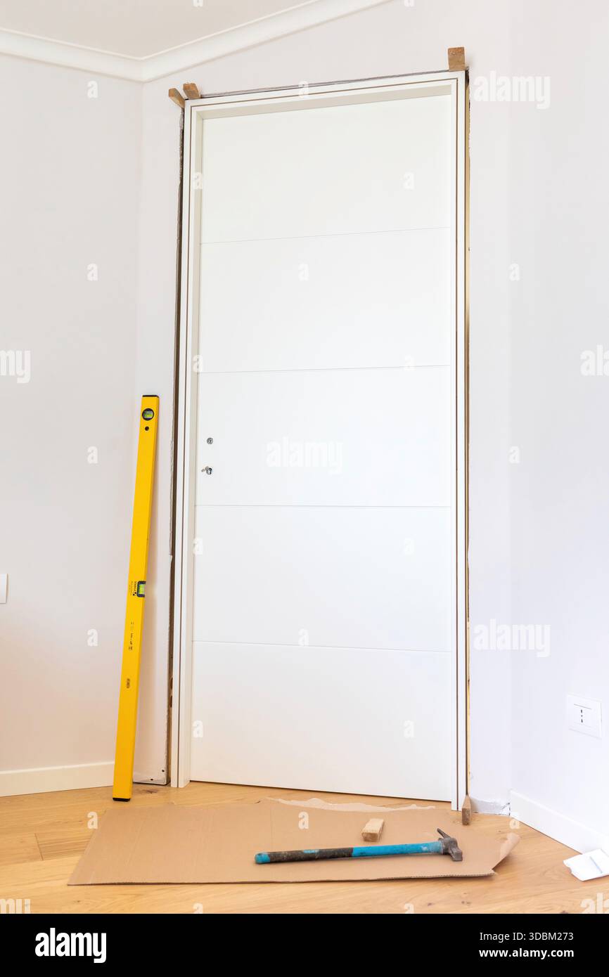 Carpenter sta installando una nuova porta interna bianca utilizzando una livella e un martello, garantendo un allineamento preciso e un posizionamento sicuro in un ambiente residenziale Foto Stock