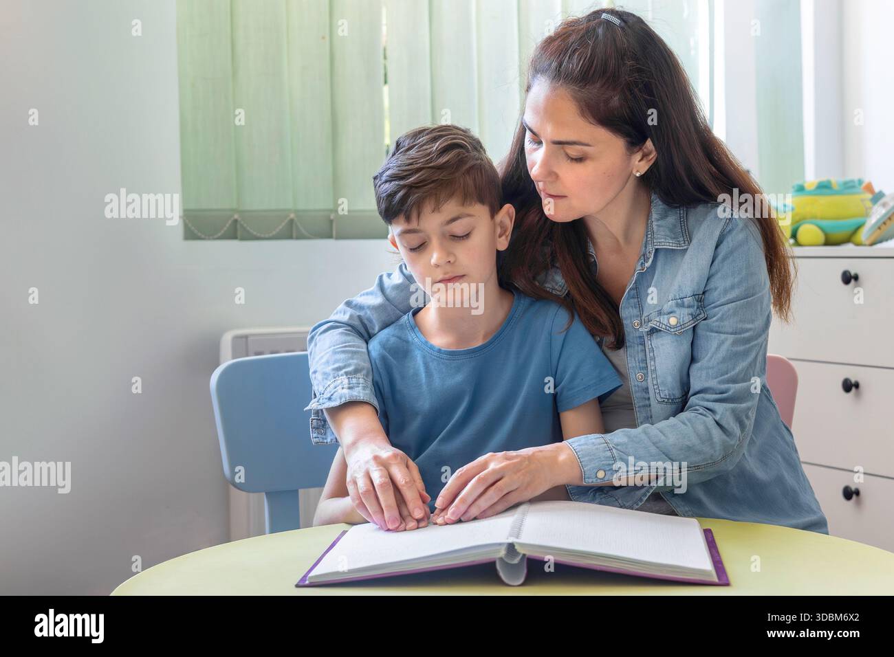 Donna che aiuta un ragazzo a leggere un libro in Braille a casa. Istruzione inclusiva e supporto all'apprendimento tattile per i bambini. Mani sul testo in Braille. Foto Stock