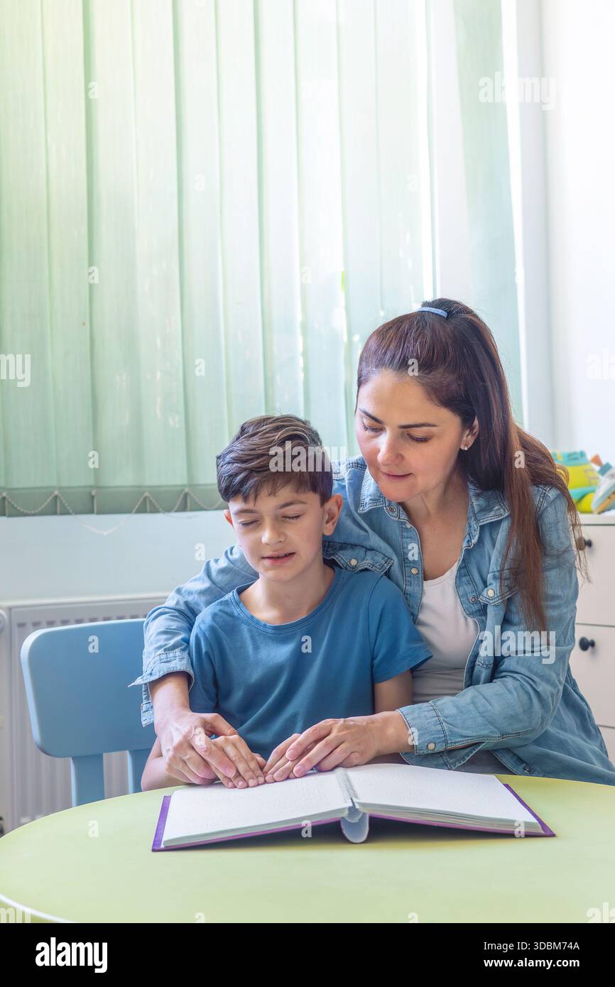 Donna che aiuta un ragazzo a leggere un libro in Braille a casa. Istruzione inclusiva e supporto all'apprendimento tattile per i bambini. Mani sul testo in Braille. Foto Stock