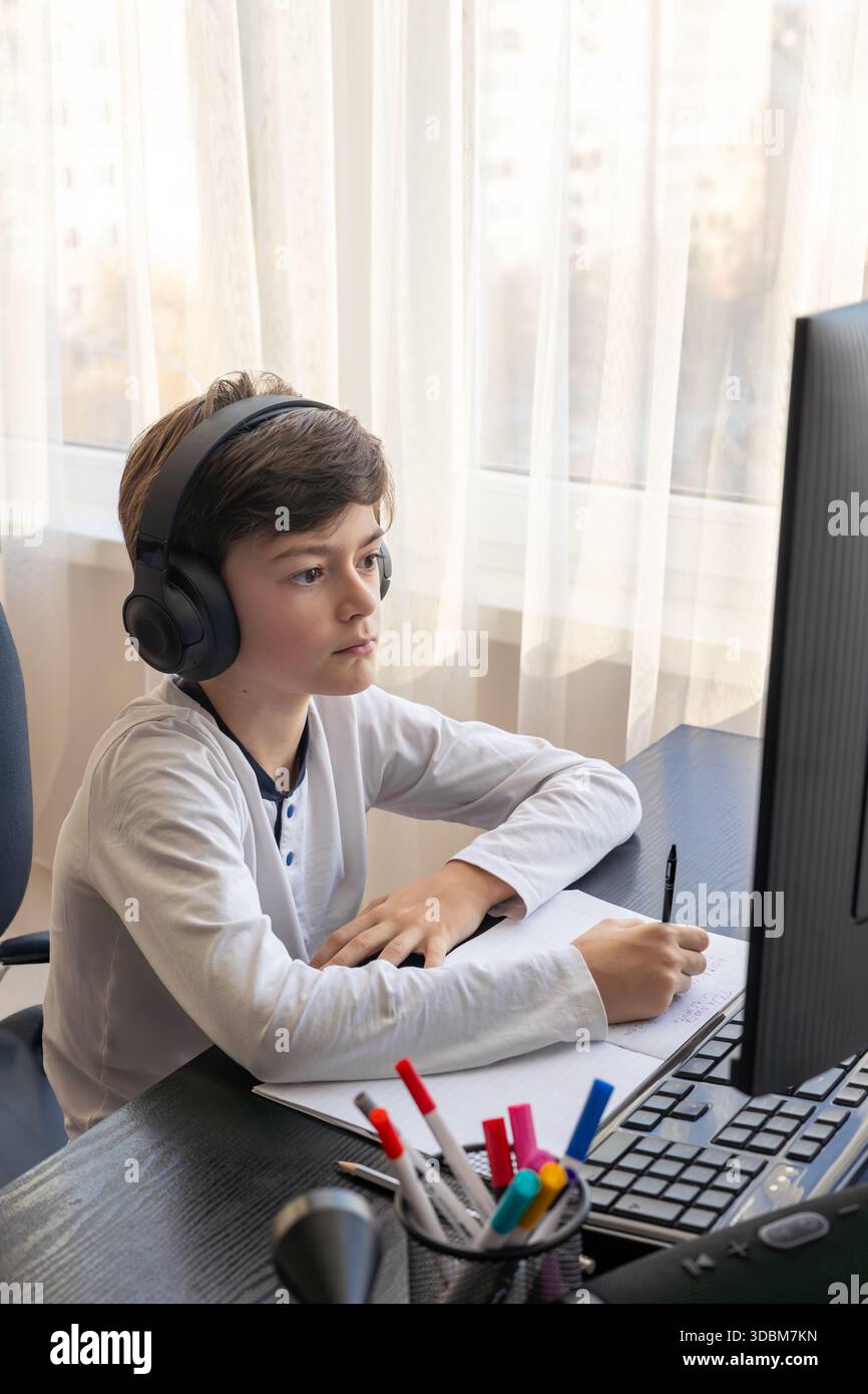 Un bambino di 7-9 anni siede a casa dietro un computer, scrivendo assegnazioni su un notebook. L'immagine acquisisce l'apprendimento remoto, le routine di studio digitali, il canale Foto Stock