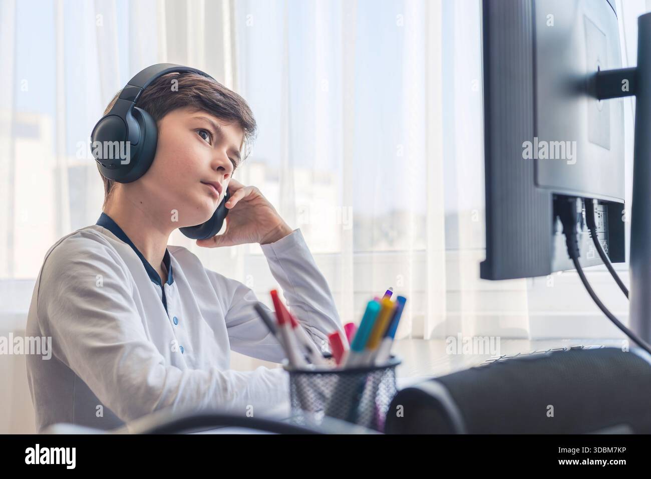 Un bambino di 7-9 anni siede a casa dietro un computer, scrivendo assegnazioni su un notebook. L'immagine acquisisce l'apprendimento remoto, le routine di studio digitali, il canale Foto Stock