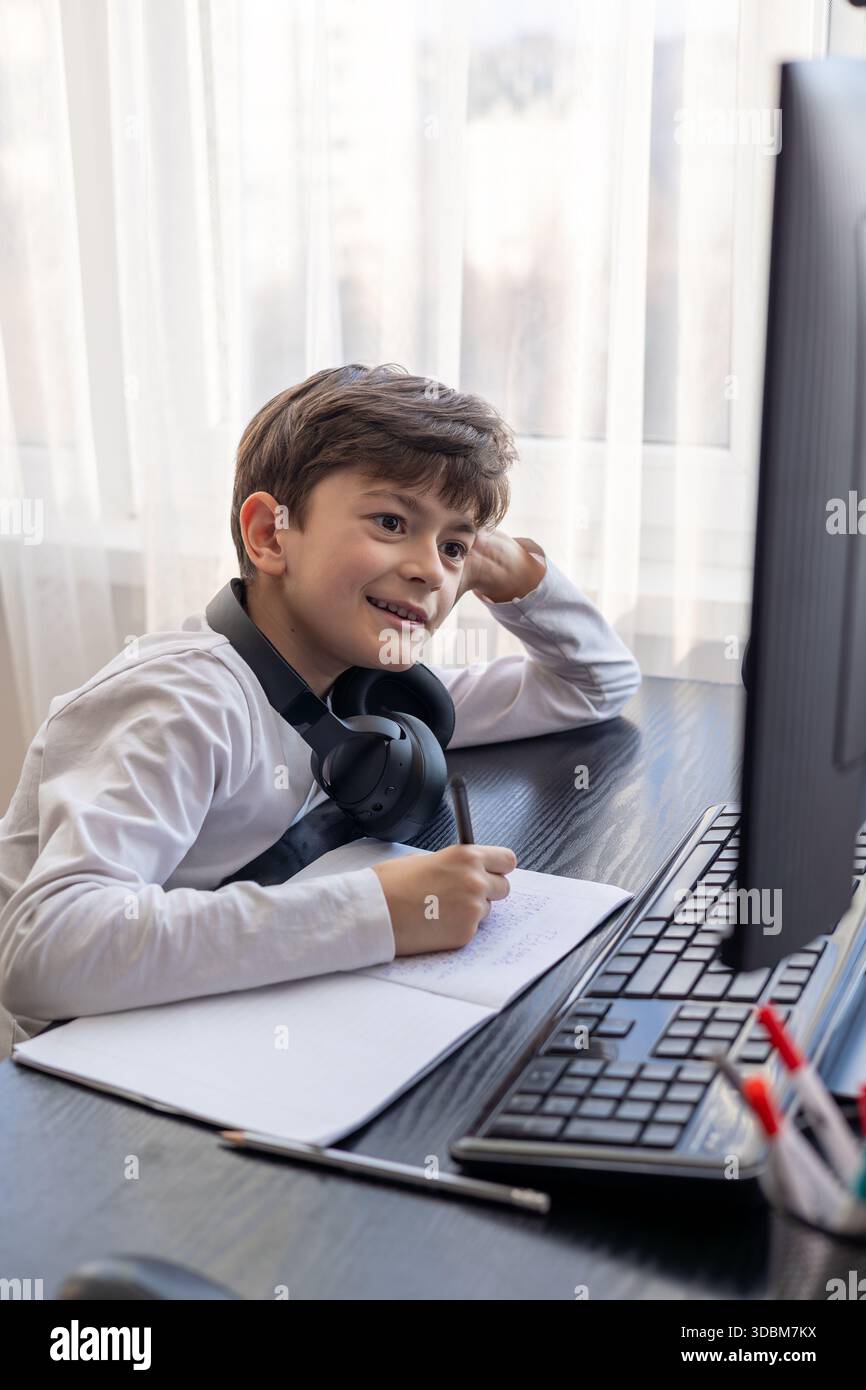 Un bambino di 7-9 anni siede a casa dietro un computer, scrivendo assegnazioni su un notebook. L'immagine acquisisce l'apprendimento remoto, le routine di studio digitali, il canale Foto Stock