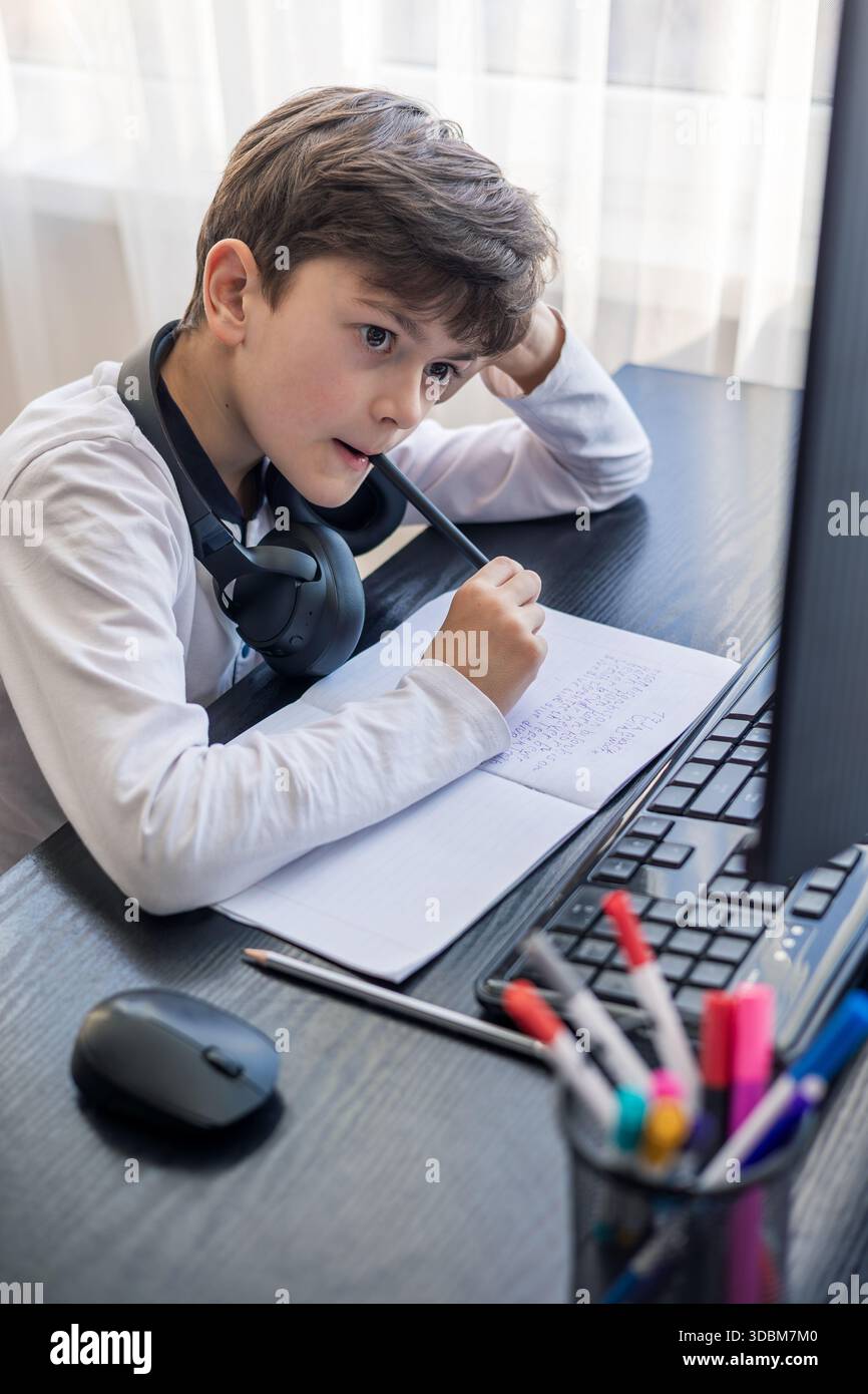 Un bambino di 7-9 anni siede a casa dietro un computer, scrivendo assegnazioni su un notebook. L'immagine acquisisce l'apprendimento remoto, le routine di studio digitali, il canale Foto Stock