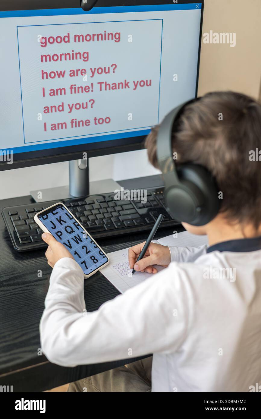 Un bambino di 7-9 anni siede a casa dietro un computer, scrivendo assegnazioni su un notebook. L'immagine acquisisce l'apprendimento remoto, le routine di studio digitali, il canale Foto Stock