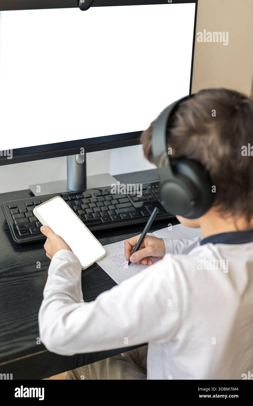 Un bambino di 7-9 anni siede a casa dietro un computer, scrivendo assegnazioni su un notebook. L'immagine acquisisce l'apprendimento remoto, le routine di studio digitali, il canale Foto Stock