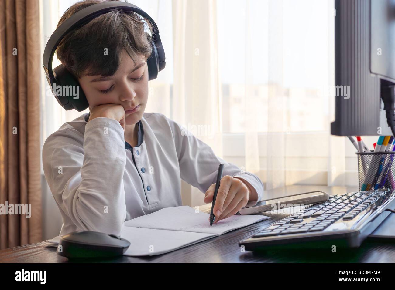 Un bambino di 7-9 anni siede a casa dietro un computer, scrivendo assegnazioni su un notebook. L'immagine acquisisce l'apprendimento remoto, le routine di studio digitali, il canale Foto Stock