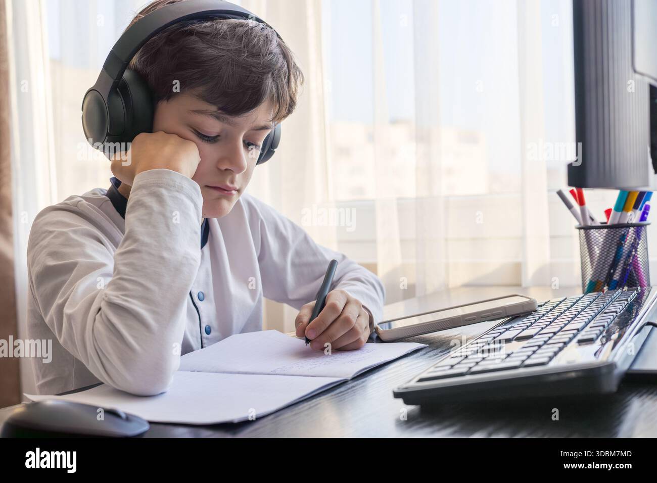 Un bambino di 7-9 anni siede a casa dietro un computer, scrivendo assegnazioni su un notebook. L'immagine acquisisce l'apprendimento remoto, le routine di studio digitali, il canale Foto Stock