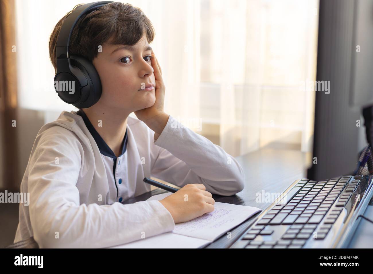 Un bambino di 7-9 anni siede a casa dietro un computer, scrivendo assegnazioni su un notebook. L'immagine acquisisce l'apprendimento remoto, le routine di studio digitali, il canale Foto Stock