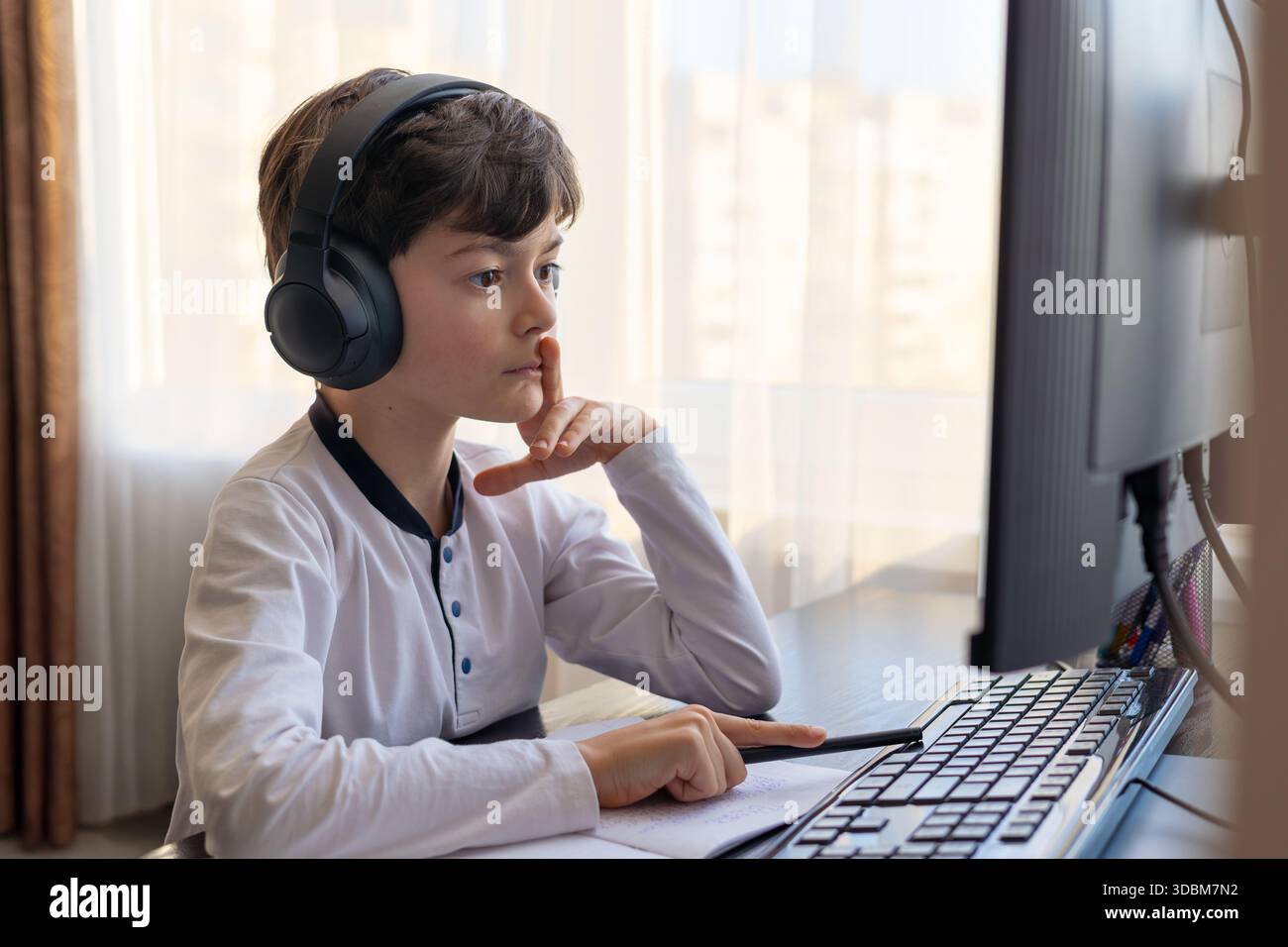 Un bambino di 7-9 anni siede a casa dietro un computer, scrivendo assegnazioni su un notebook. L'immagine acquisisce l'apprendimento remoto, le routine di studio digitali, il canale Foto Stock