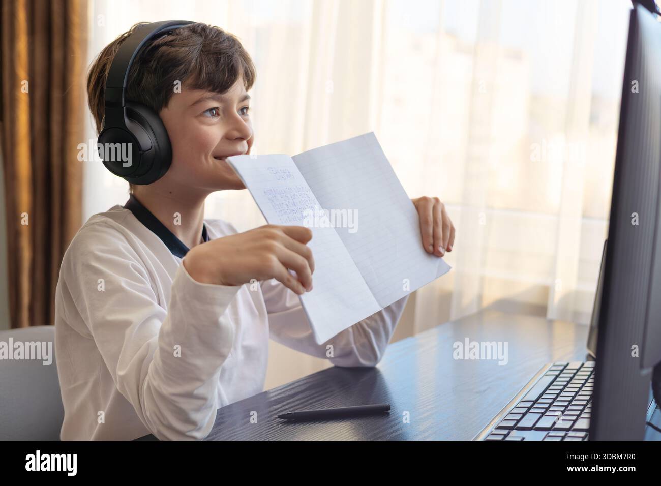 Un ragazzo sorridente di 7-9 anni studia online alla sua scrivania. Mostra con orgoglio il suo quaderno di compiti sullo schermo, catturando l'apprendimento a casa e gli educati a distanza Foto Stock