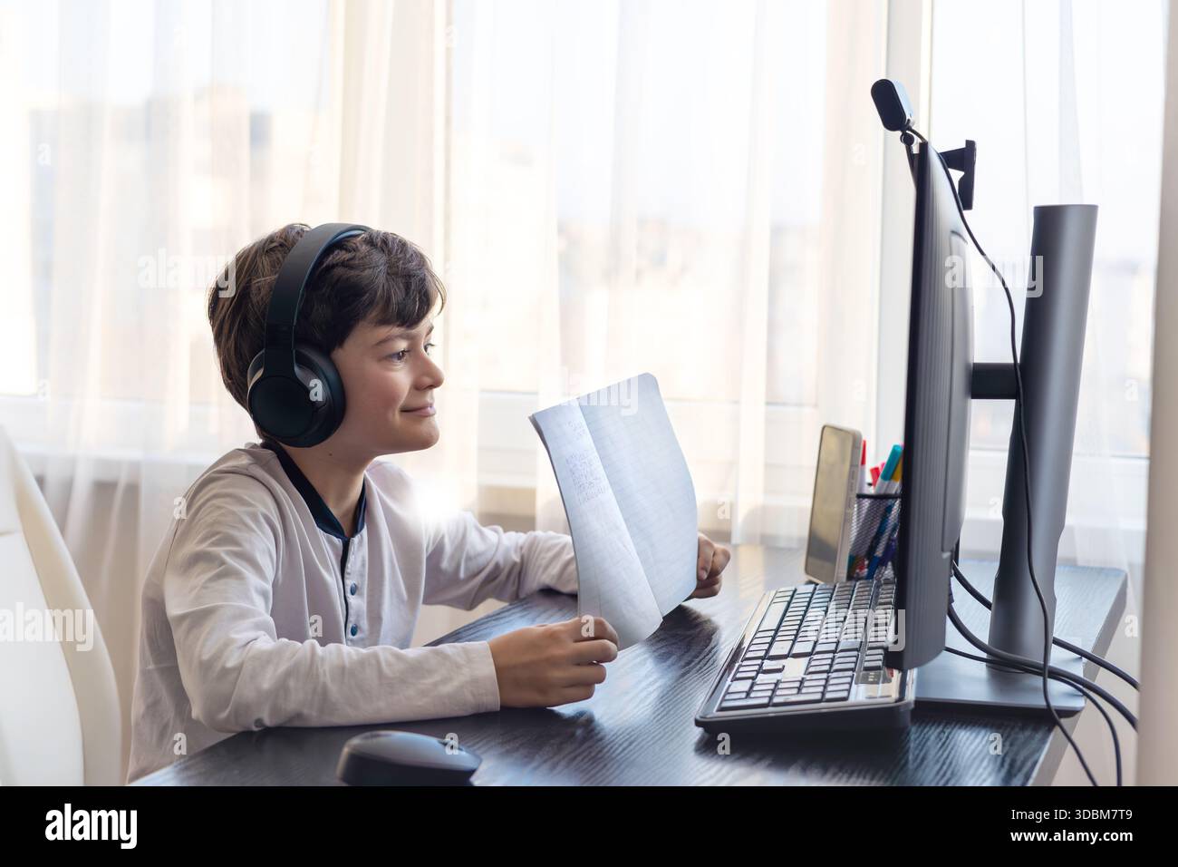 Un ragazzo sorridente di 7-9 anni studia online alla sua scrivania. Mostra con orgoglio il suo quaderno di compiti sullo schermo, catturando l'apprendimento a casa e gli educati a distanza Foto Stock