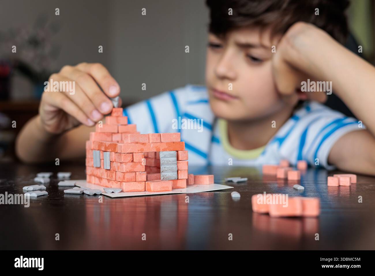 Triste ragazzo seduto a casa, sembra annoiato e non motivato mentre cerca di costruire una casa di mattoni giocattolo. Emozioni infantili, difficoltà di apprendimento, mancanza di foc Foto Stock