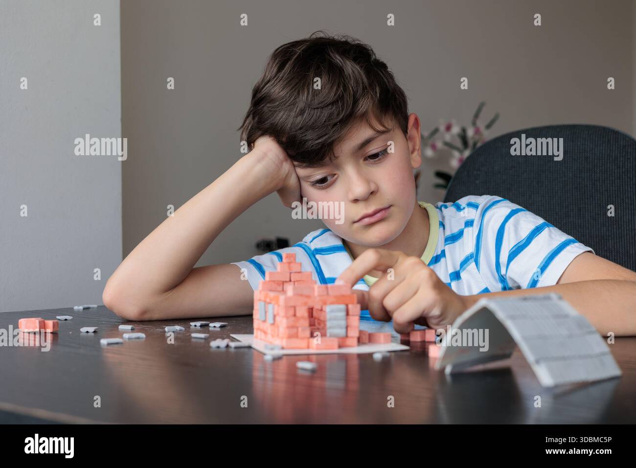 Triste ragazzo seduto a casa, sembra annoiato e non motivato mentre cerca di costruire una casa di mattoni giocattolo. Emozioni infantili, difficoltà di apprendimento, mancanza di foc Foto Stock