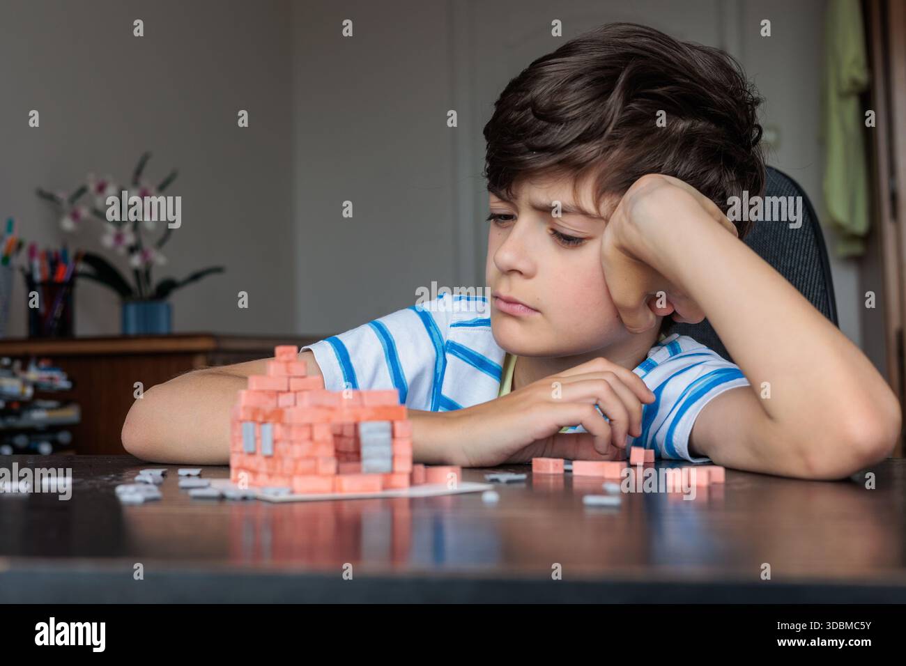 Triste ragazzo seduto a casa, sembra annoiato e non motivato mentre cerca di costruire una casa di mattoni giocattolo. Emozioni infantili, difficoltà di apprendimento, mancanza di foc Foto Stock
