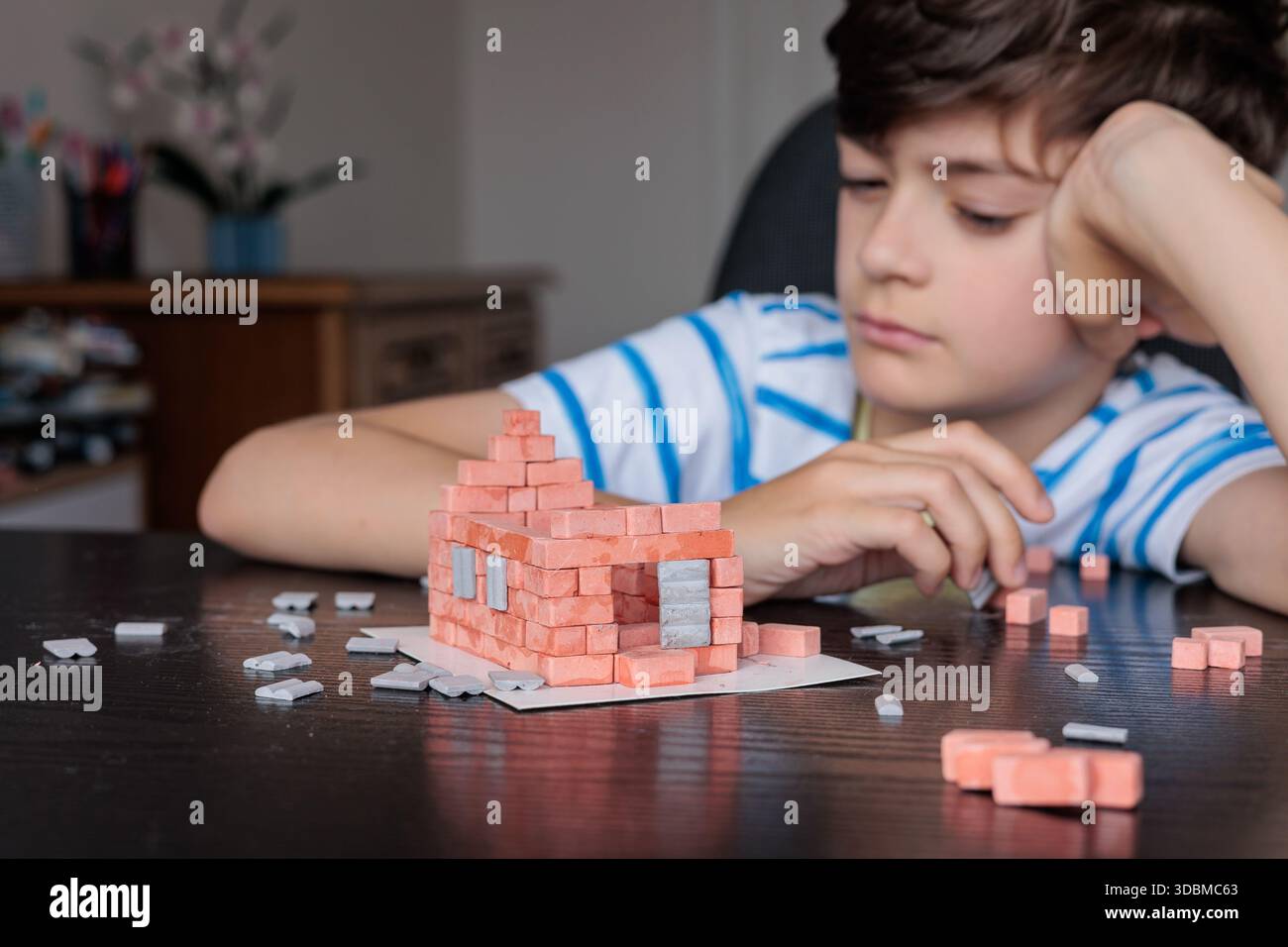 Triste ragazzo seduto a casa, sembra annoiato e non motivato mentre cerca di costruire una casa di mattoni giocattolo. Emozioni infantili, difficoltà di apprendimento, mancanza di foc Foto Stock