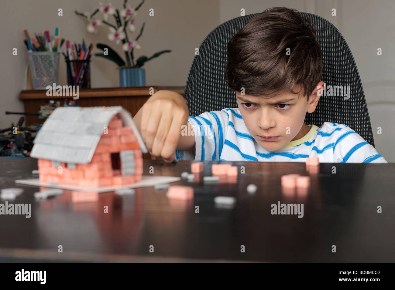Triste ragazzo seduto a casa, sembra annoiato e non motivato mentre cerca di costruire una casa di mattoni giocattolo. Emozioni infantili, difficoltà di apprendimento, mancanza di foc Foto Stock
