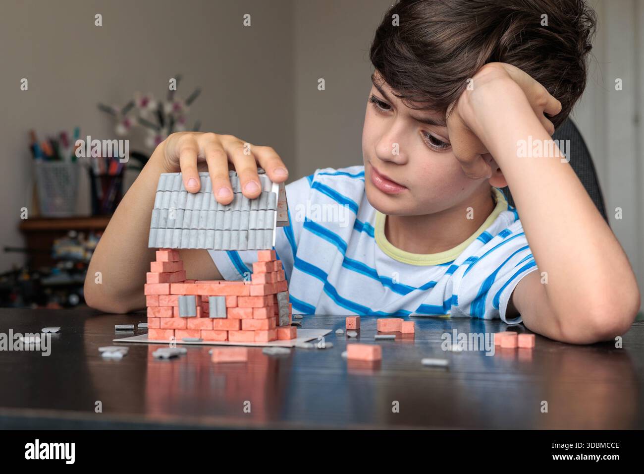 Triste ragazzo seduto a casa, sembra annoiato e non motivato mentre cerca di costruire una casa di mattoni giocattolo. Emozioni infantili, difficoltà di apprendimento, mancanza di foc Foto Stock