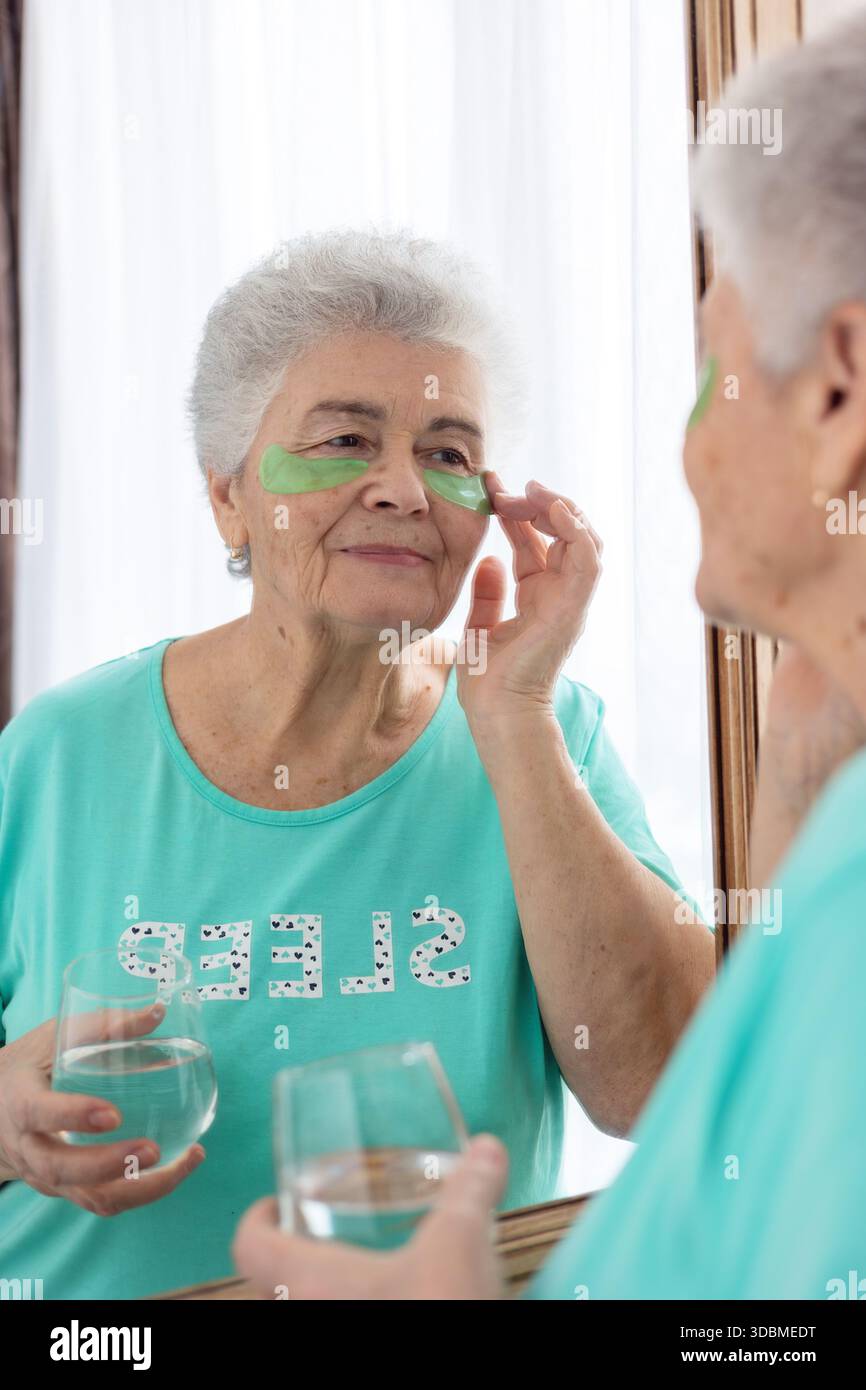 Donna anziana che si prende cura della sua pelle, applica cerotti sotto gli occhi, guardando il suo riflesso nello specchio. Un momento di autostima e bellezza, concentrandosi su Foto Stock