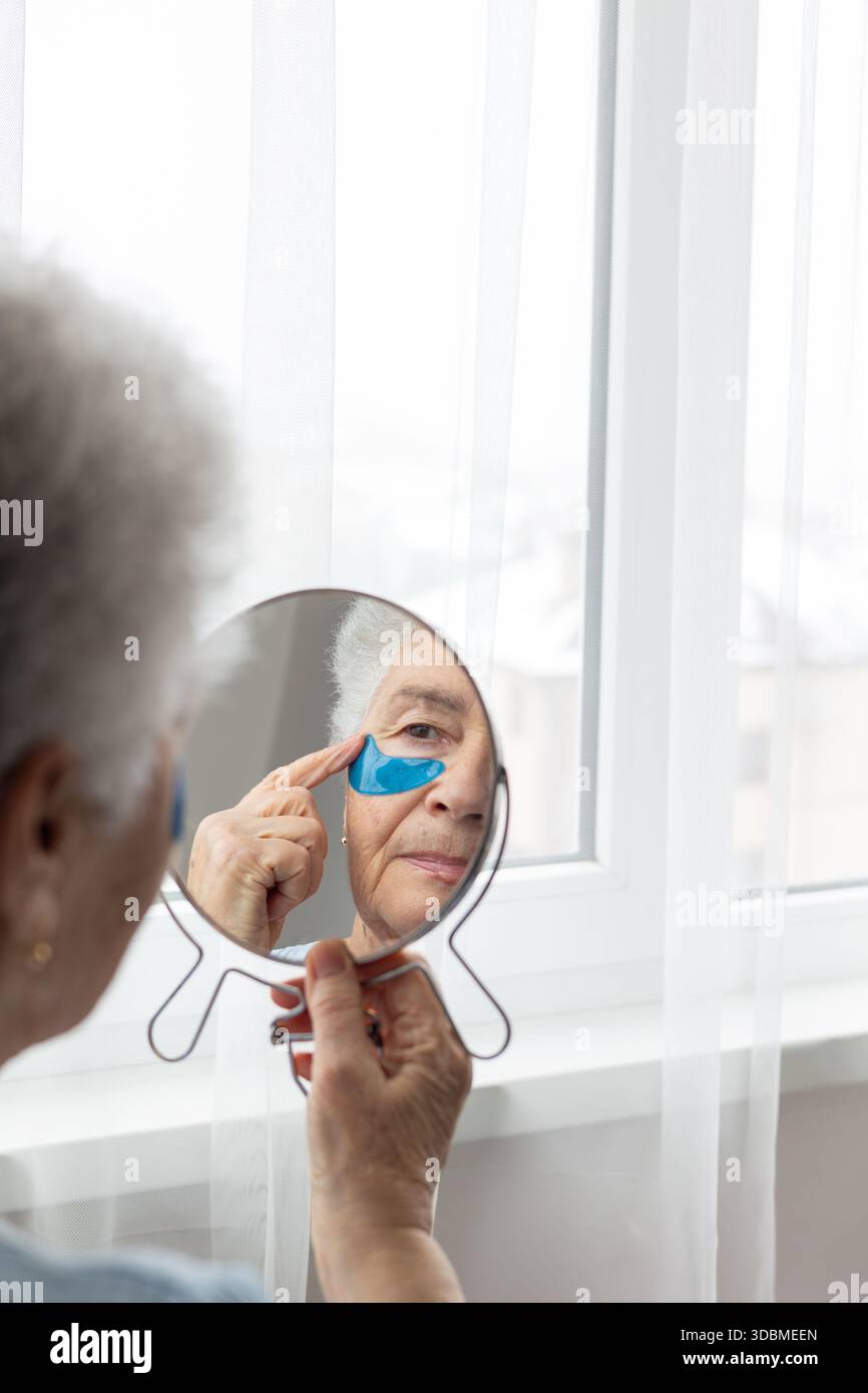 Donna anziana che si prende cura della sua pelle, applica cerotti sotto gli occhi, guardando il suo riflesso nello specchio. Un momento di autostima e bellezza, concentrandosi su Foto Stock