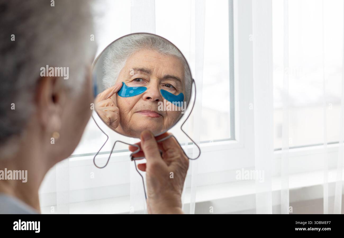 Donna anziana che si prende cura della sua pelle, applica cerotti sotto gli occhi, guardando il suo riflesso nello specchio. Un momento di autostima e bellezza, concentrandosi su Foto Stock