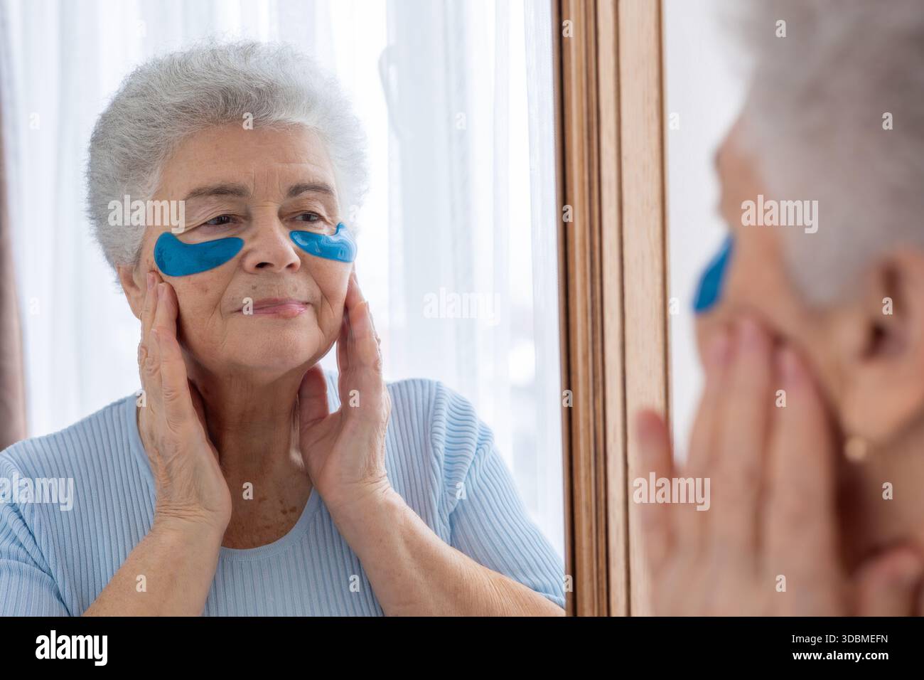 Donna anziana che si prende cura della sua pelle, applica cerotti sotto gli occhi, guardando il suo riflesso nello specchio. Un momento di autostima e bellezza, concentrandosi su Foto Stock