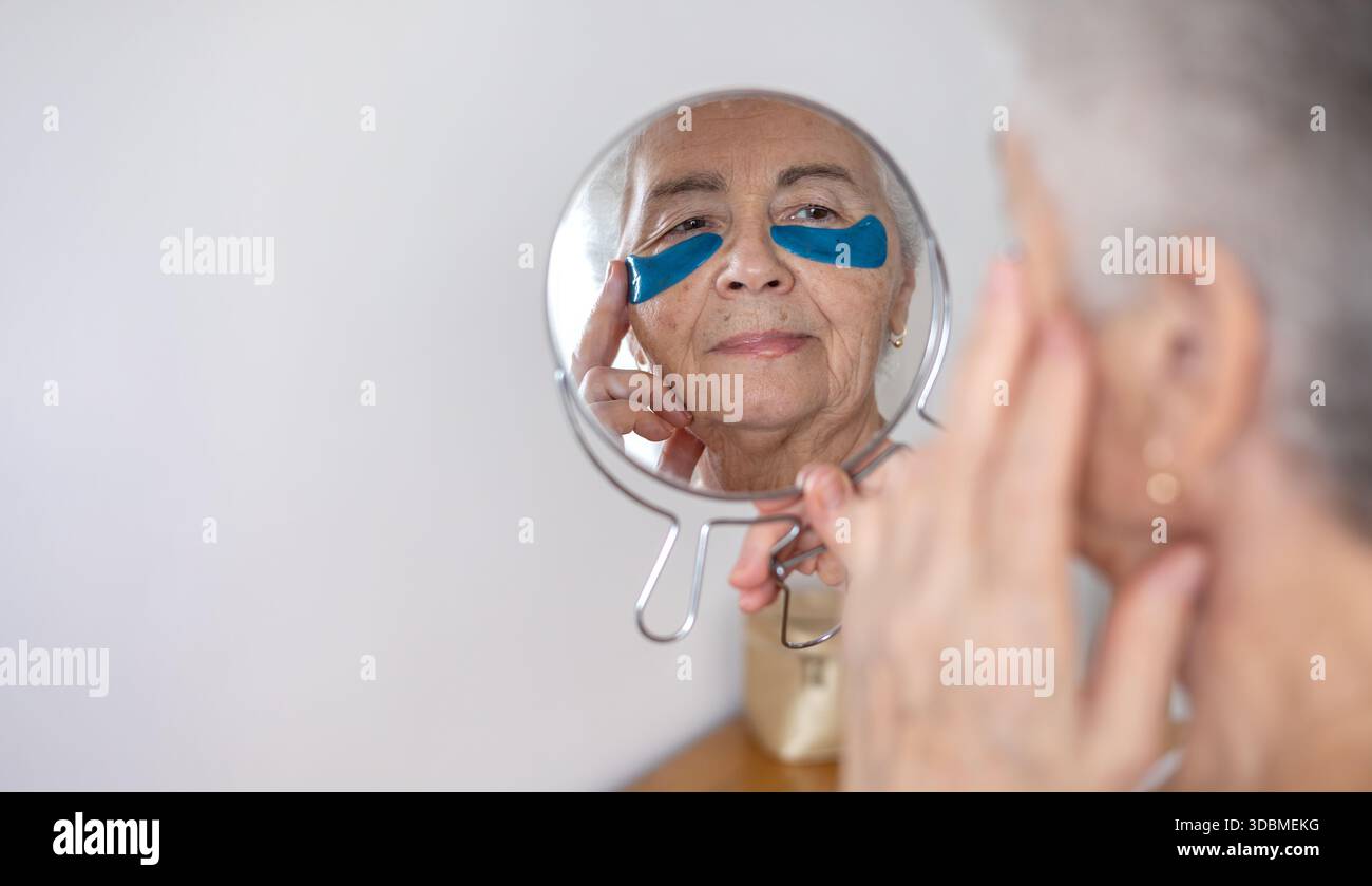 Donna anziana che si prende cura della sua pelle, applica cerotti sotto gli occhi, guardando il suo riflesso nello specchio. Un momento di autostima e bellezza, concentrandosi su Foto Stock