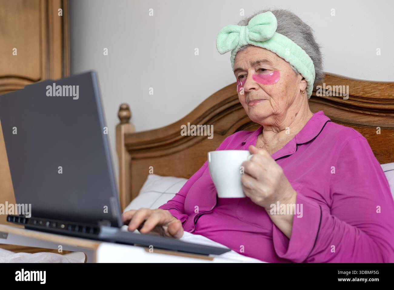Donna anziana di oltre 70 anni in toppe per gli occhi che si rilassano a letto con un laptop e un tè, godendosi l'autoassistenza mattutina e la connessione digitale. Copia spazio Foto Stock