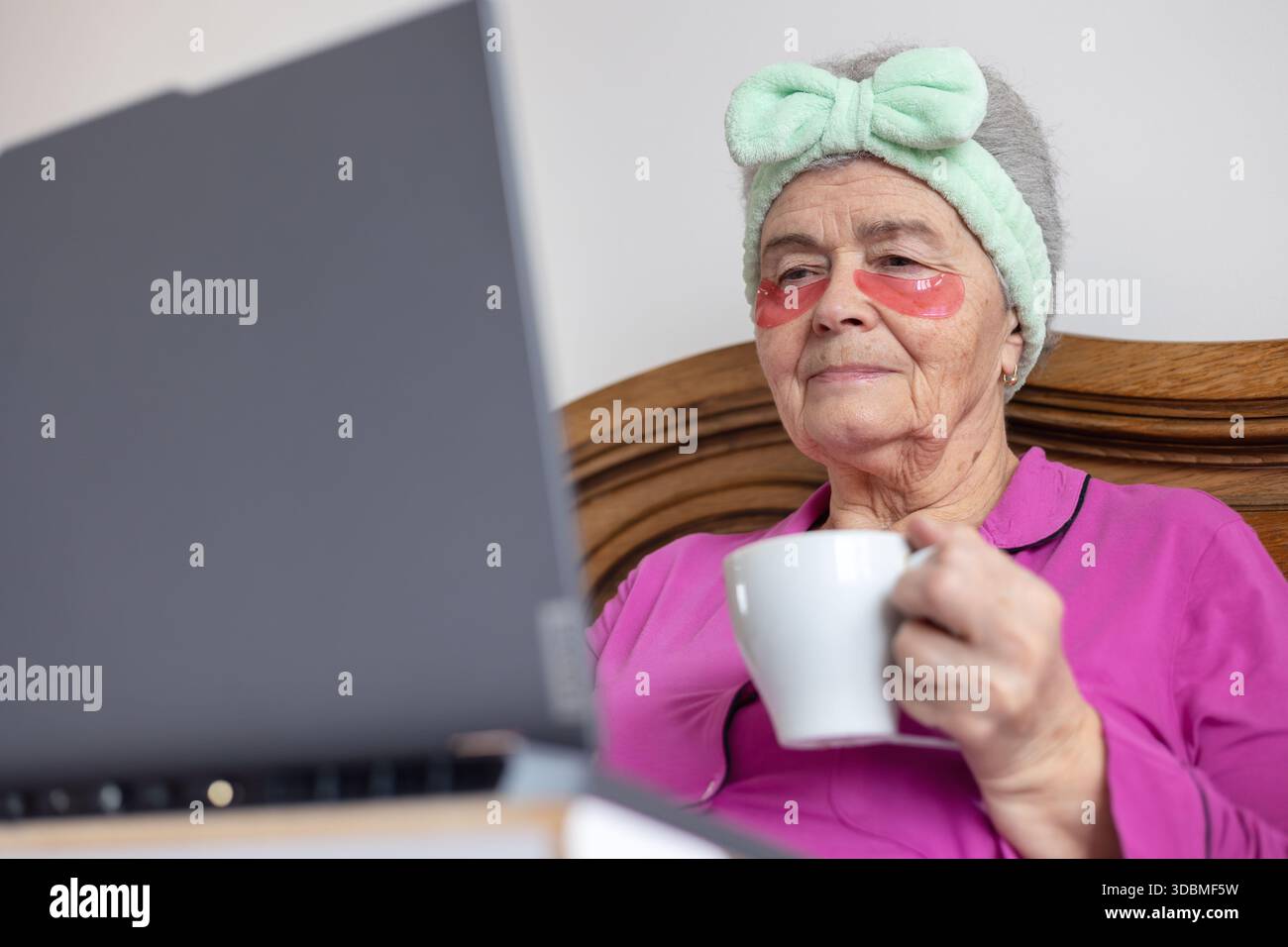 Donna anziana di oltre 70 anni in toppe per gli occhi che si rilassano a letto con un laptop e un tè, godendosi l'autoassistenza mattutina e la connessione digitale. Copia spazio Foto Stock