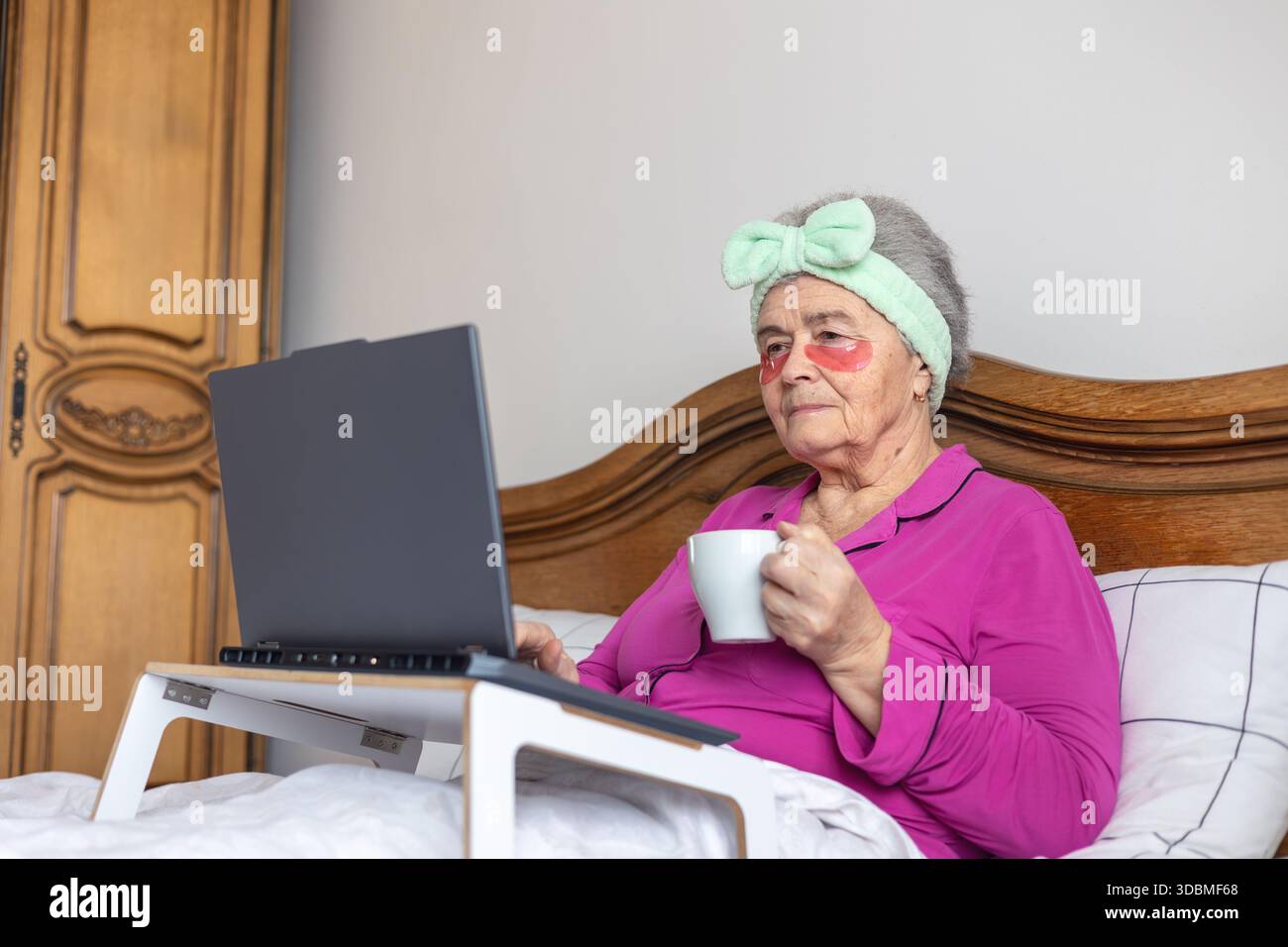Donna anziana di oltre 70 anni in toppe per gli occhi che si rilassano a letto con un laptop e un tè, godendosi l'autoassistenza mattutina e la connessione digitale. Copia spazio Foto Stock