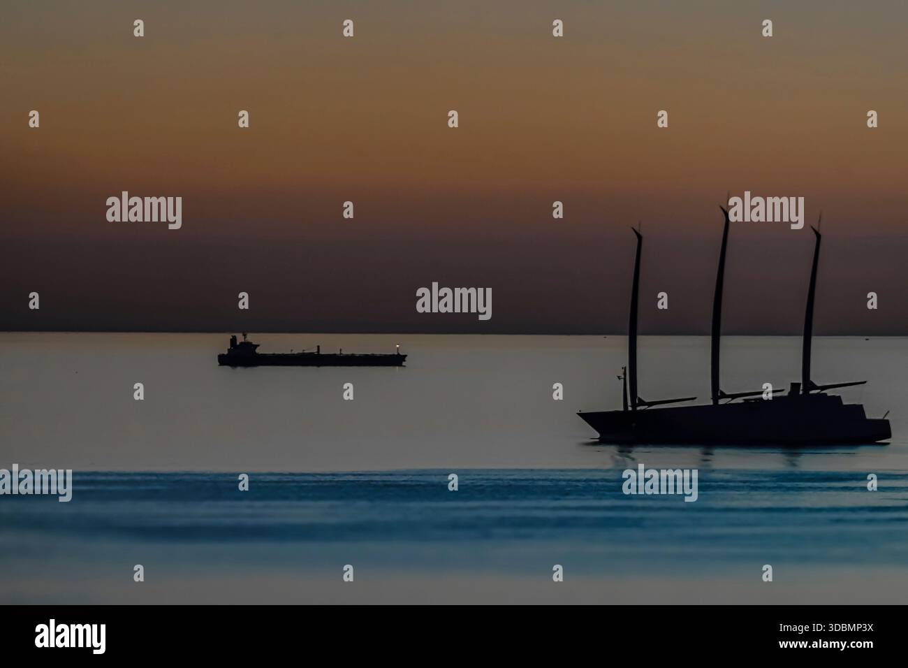 Trieste, Italia, vista mare serale con sagome di due navi in acqua calma, uno yacht a vela con tre alberi e una nave da carico in lontananza sotto un cielo in pendenza al crepuscolo? [M] Foto Stock
