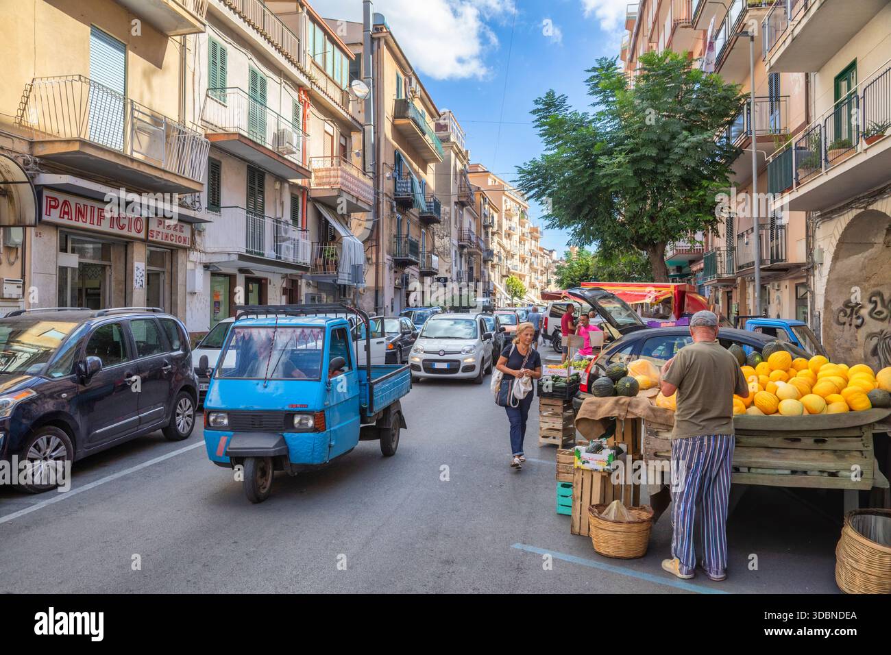 Mercato di strada a Monreale, in Sicilia, con la gente del posto, prodotti freschi in un mercato all'aperto, mentre un classico veicolo Ape Piaggio attraversa la strada. Monreale, Palermo, Sicilia, Italia Foto Stock