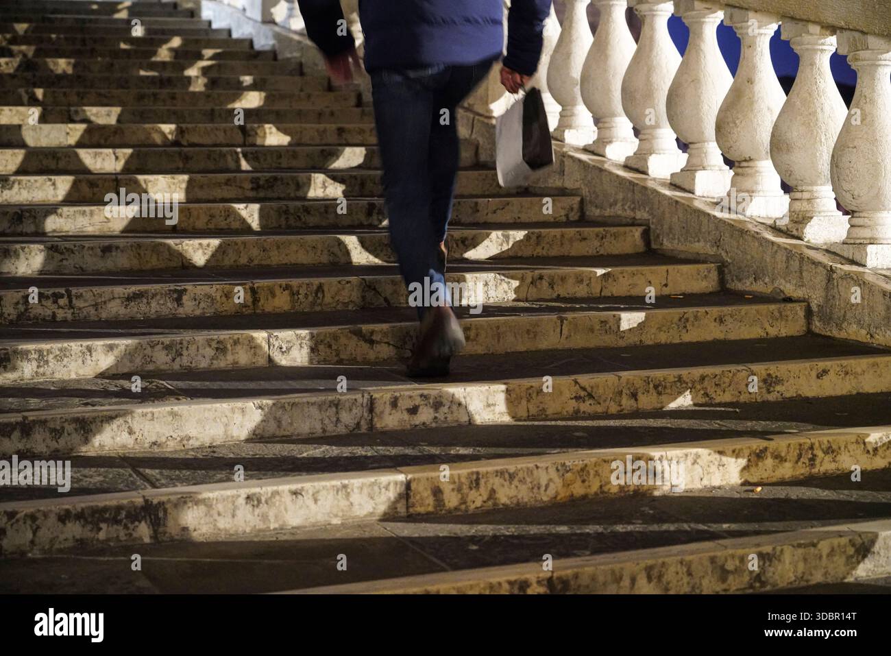 Un uomo cammina sui gradini del Ponte di Rialto a Venezia, in Italia, di notte, portando una borsa della spesa vicino a una balaustra di pietra bianca. Foto Stock