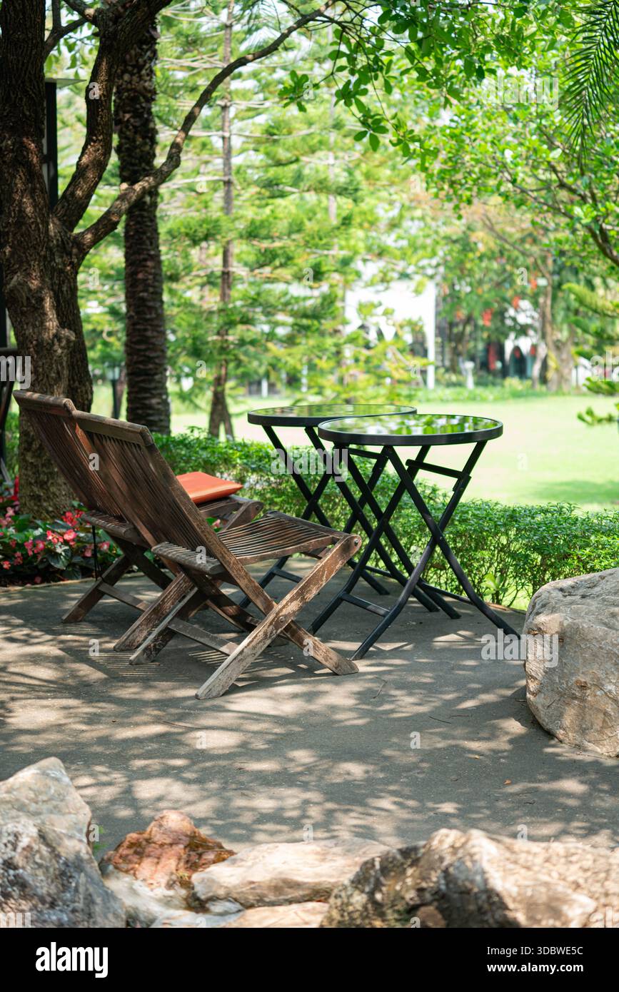 Rilassante area salotto all'aperto in un lussureggiante giardino verde. Presenta sedie rustiche in legno e tavoli rotondi in vetro all'ombra degli alberi con una bella na Foto Stock