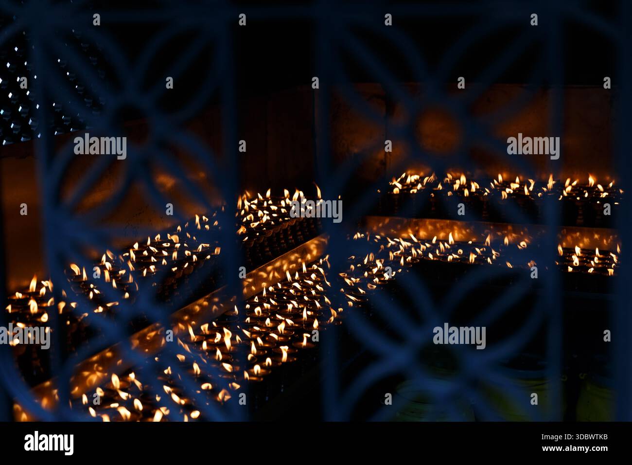 File di lampade a olio luccicanti del tempio viste attraverso una griglia in metallo decorativo, creando uno sfondo astratto di calda luce del fuoco e atmosfera spirituale. Foto Stock