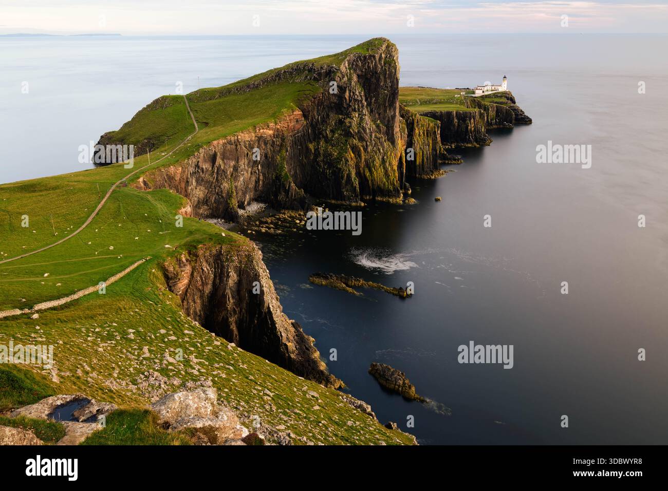 Neist Point con un piccolo faro posizionato alla fine di una massiccia scogliera verticale con prati verdi e mare d'argento calmo leggermente alleggerito dal soffice wa Foto Stock