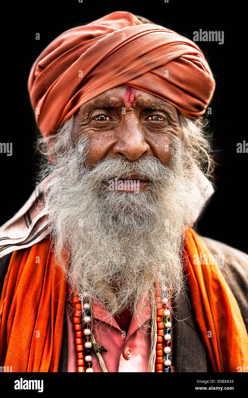 Un ritratto di un uomo anziano, forse un Sadhu o un anziano del villaggio indiano, che indossa un turbante arancione, una sciarpa e una lunga barba grigia contro un bac nero Foto Stock