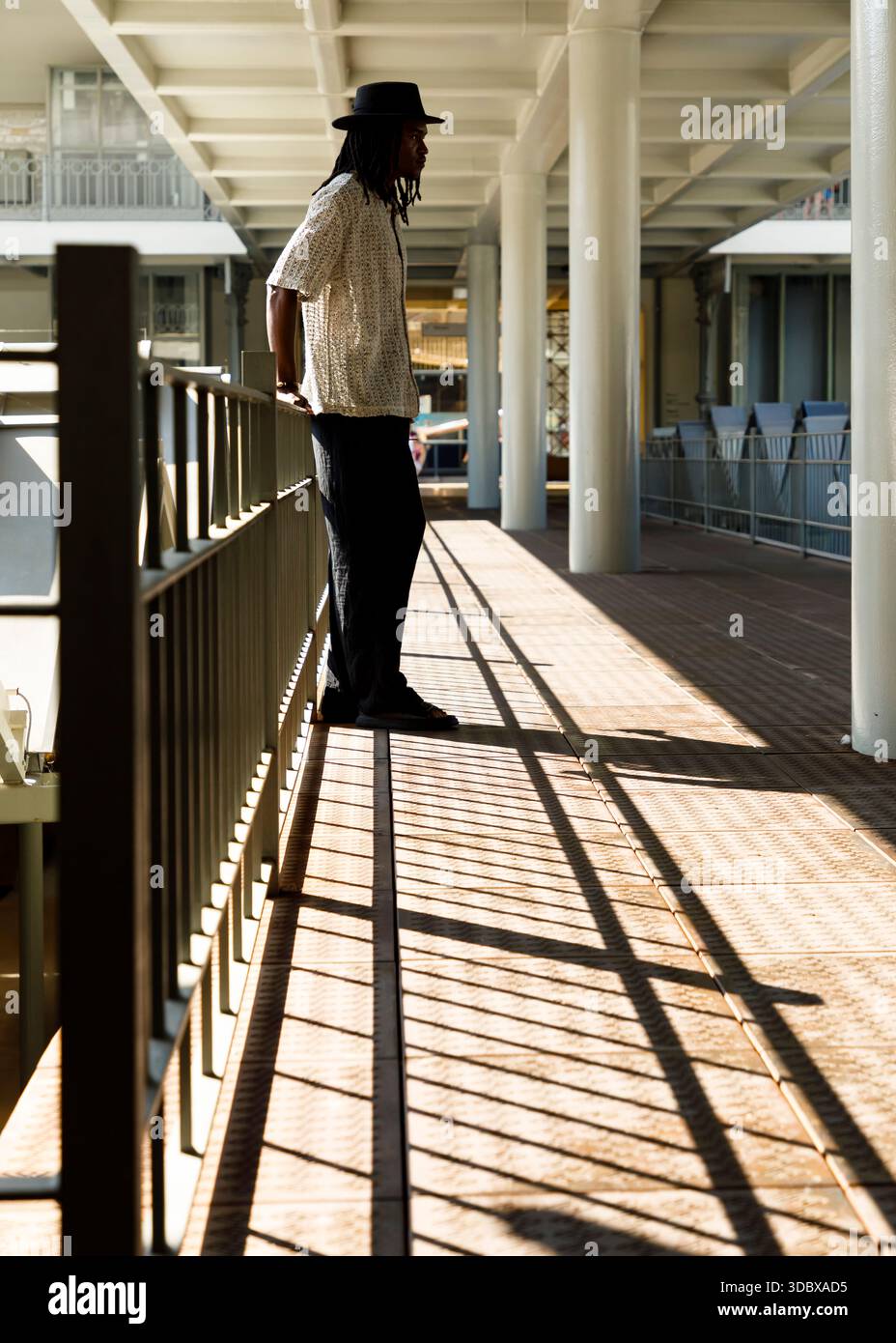 Un uomo che indossa un cappello e una camicia a motivi geometrici si erge su un passaggio pedonale, con ombre sparse per terra. Sullo sfondo, due donne stanno in piedi e parlano Foto Stock