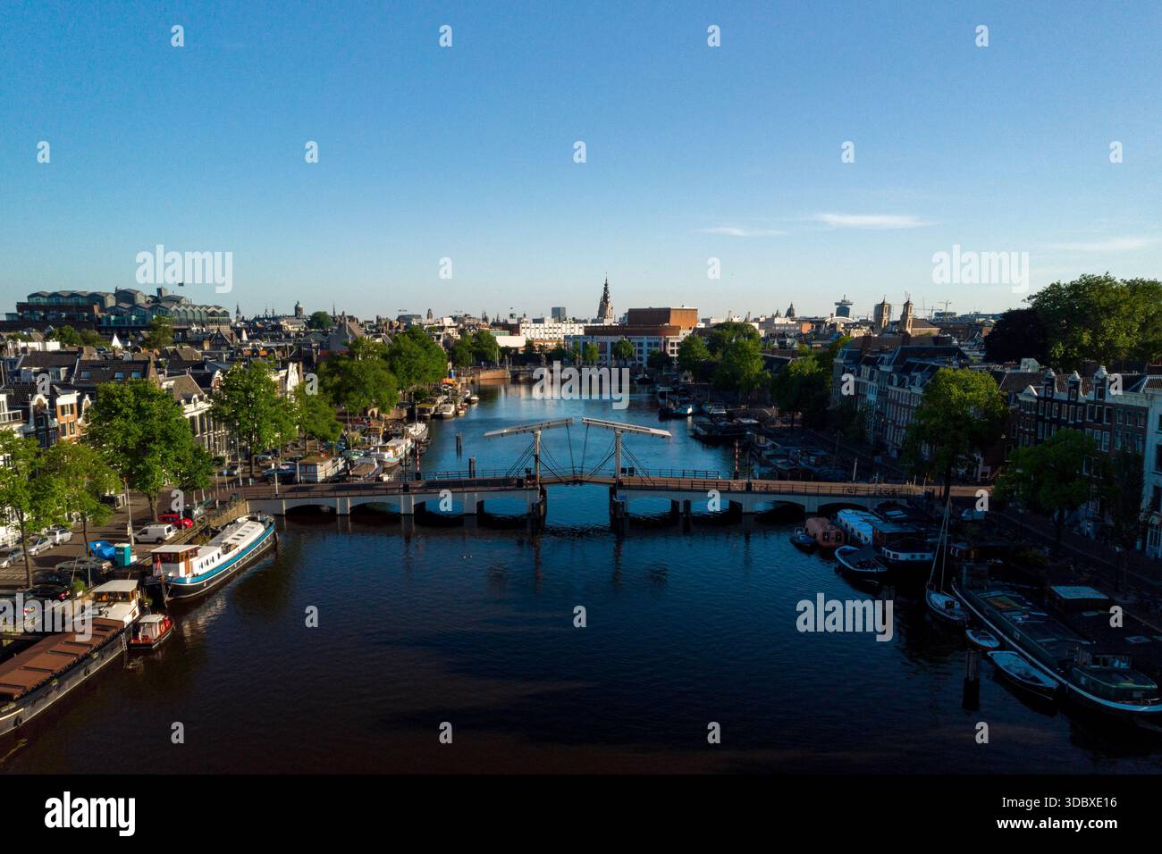 Vista aerea delle acque scure dell'Amstel che riflettono il paesaggio urbano e il Ponte Magere, incorniciato da alberi lussureggianti sotto un cielo limpido, Amsterdam, North Holl Foto Stock