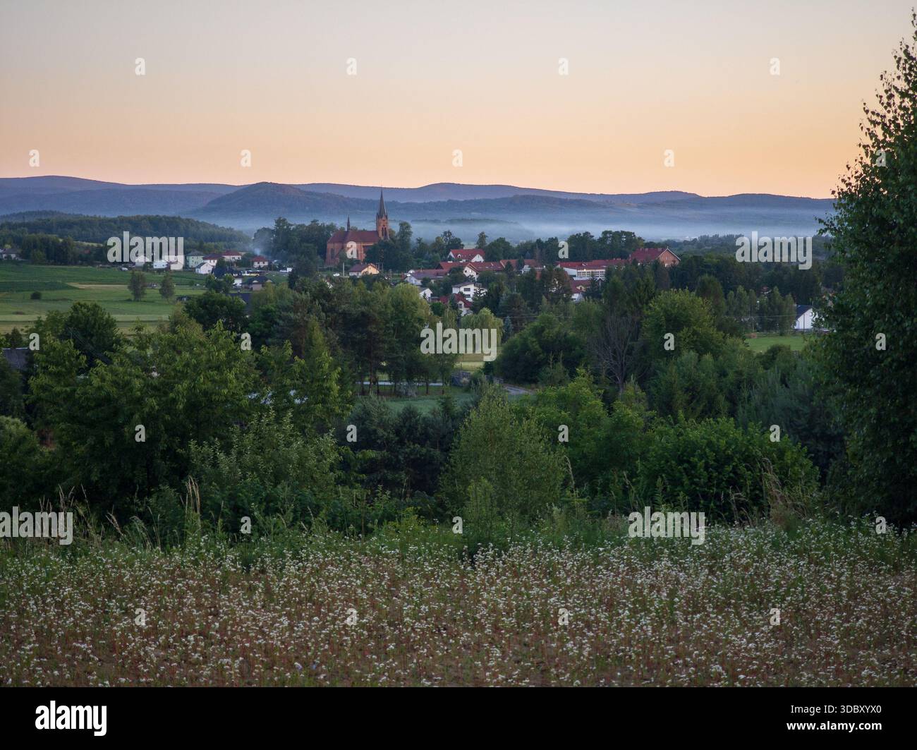 Crepuscolo nebbioso sopra il villaggio di Osobnica in Polonia con la chiesa neogotica di Talowski che si innalza sopra la valle contro il Monte Low Beskid. Foto Stock