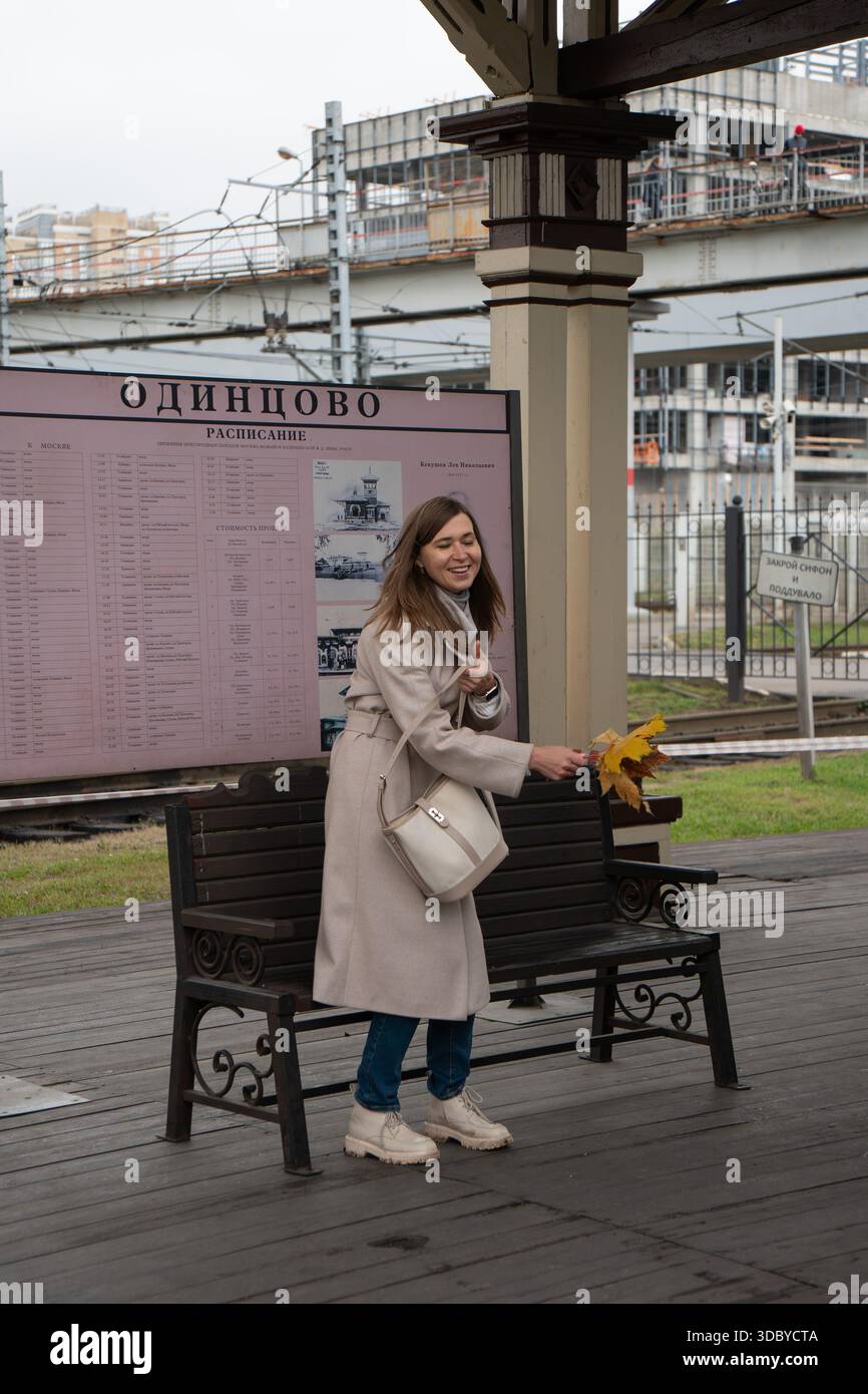 Mosca, Russia - 18 ottobre 2025: Donna in piedi vicino alla panchina della stazione ferroviaria, sorridendo e tenendo le foglie d'autunno davanti agli orari di Odintsovo Foto Stock