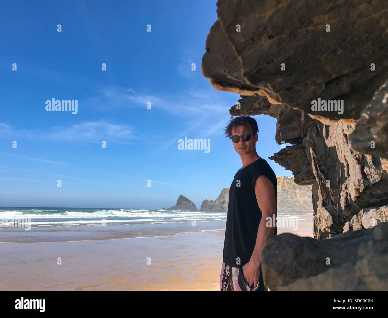 Bell'uomo in un elegante abito estivo all'ombra della roccia, godendosi il selvaggio paesaggio della spiaggia a Praia da Amoreira, Portogallo. Viaggi, moda. Foto Stock