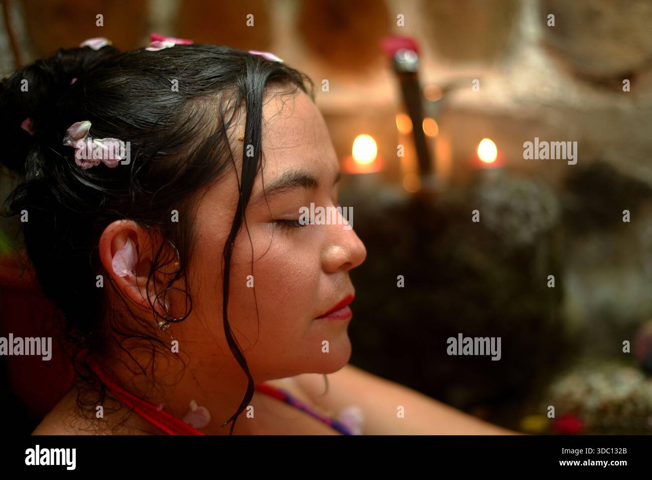 Donna che trova pace e calma durante un rilassante bagno termale, i suoi capelli bagnati sono decorati con petali di fiori mentre le candele brillano sullo sfondo, simboleggiando la Foto Stock