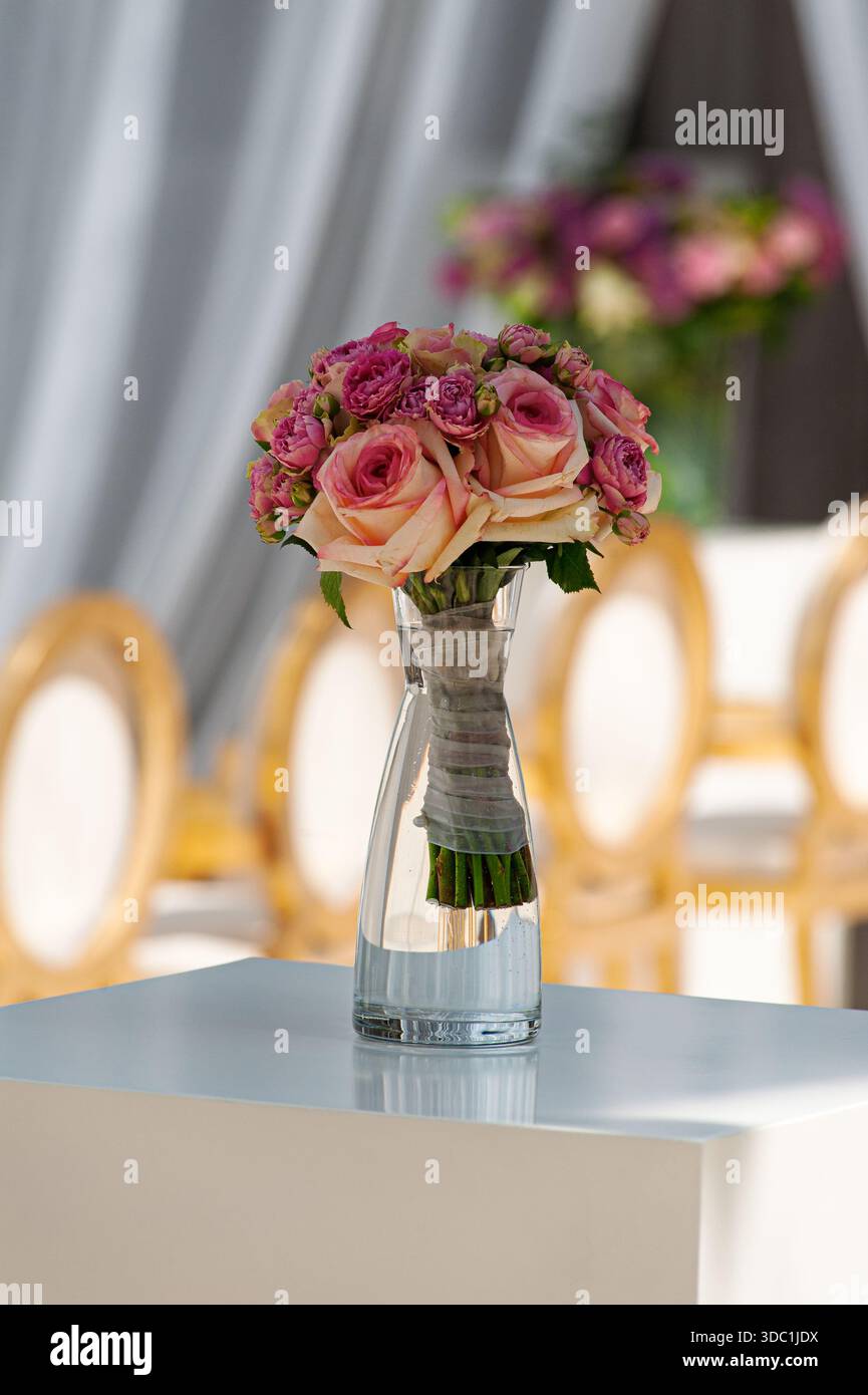 Un romantico bouquet di rose rosa disposto come pezzo centrale in un vaso di vetro trasparente, avvolto con spago. Gli arredi morbidi ed eleganti creano un matrimonio raffinato o una festa speciale Foto Stock