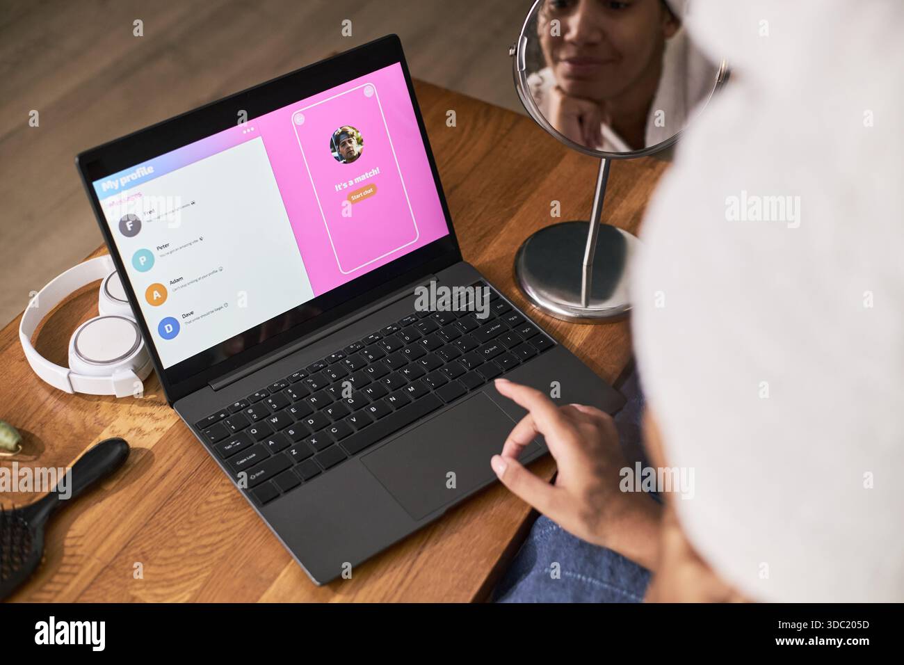 Giovane donna nera adulta che usa il laptop per incontri online, tocca il trackpad mentre guarda il profilo degli incontri sullo schermo, riflesso del viso visibile nello specchio rotondo sulla scrivania Foto Stock
