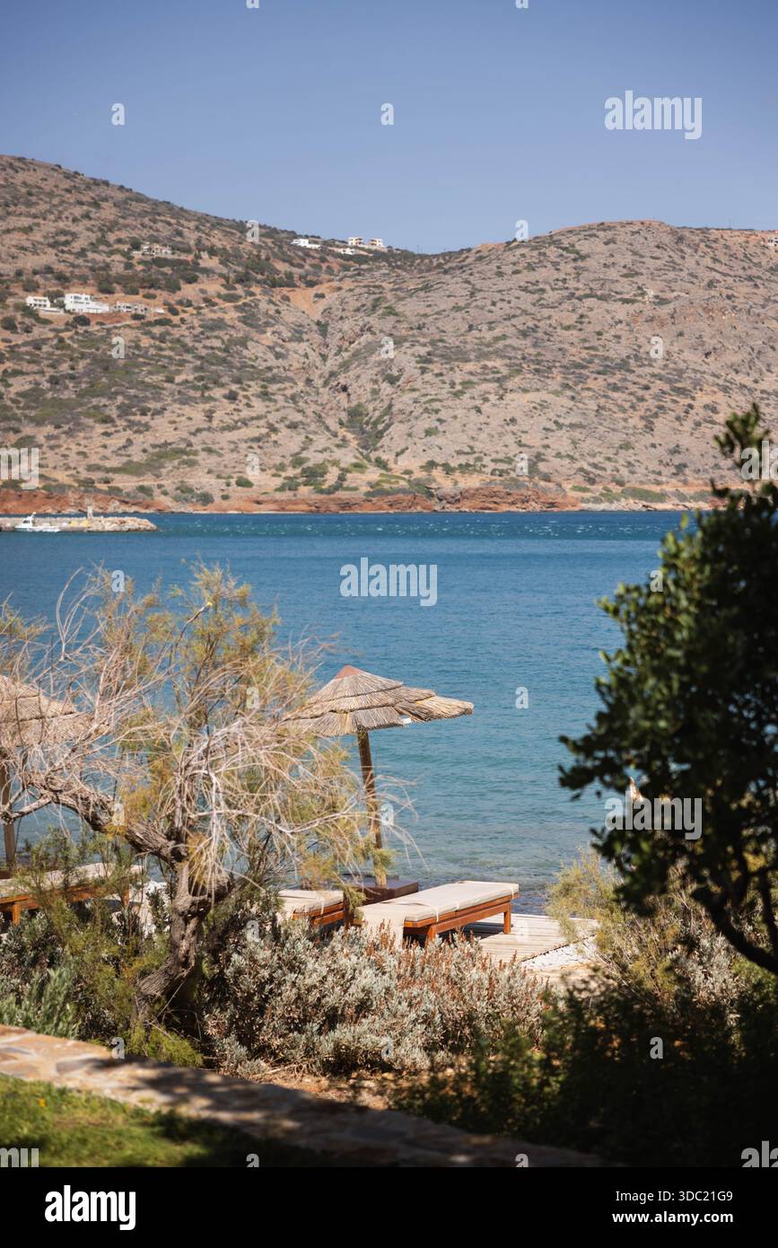 Resort sulla spiaggia con lettini e ombrellone che si affaccia sulle acque turchesi del Mediterraneo e sulle colline aride, Creta Grecia. Paradiso delle vacanze nelle isole greche Foto Stock