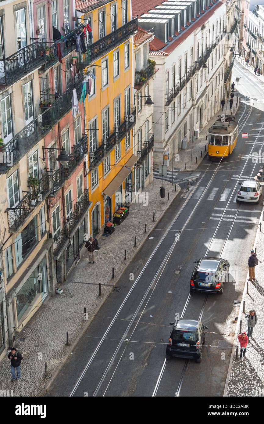 Vista dall'alto dell'iconico tram giallo 28 che attraversa una stradina stretta di Lisbona, Portogallo, circondata da edifici colorati e automobili. Foto Stock