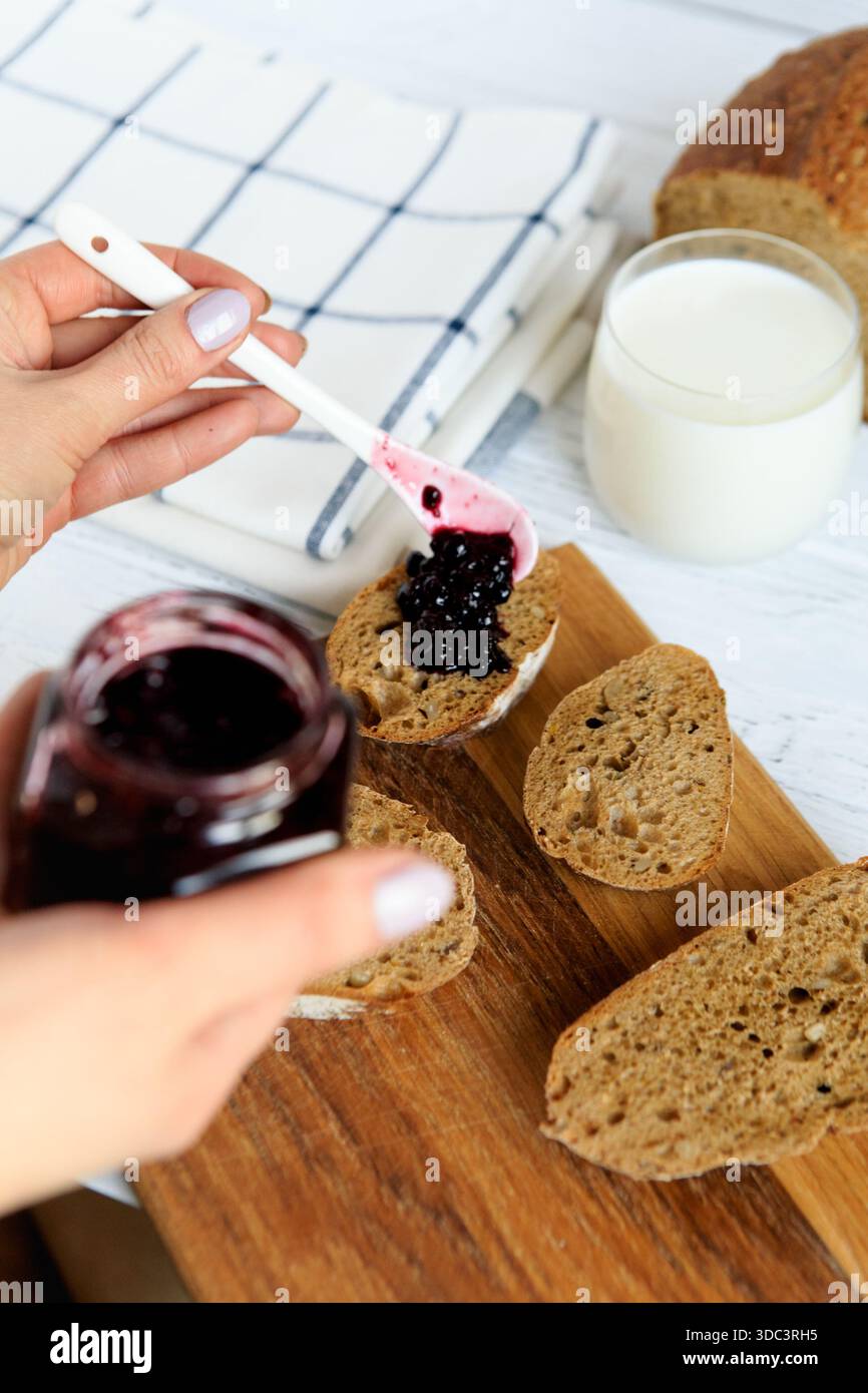 Fette di pane fatto in casa condite con marmellata di bacche scure su una tavola di legno, servite con latte. Luminosa cucina arredata in un ambiente accogliente e casalingo per la colazione Foto Stock
