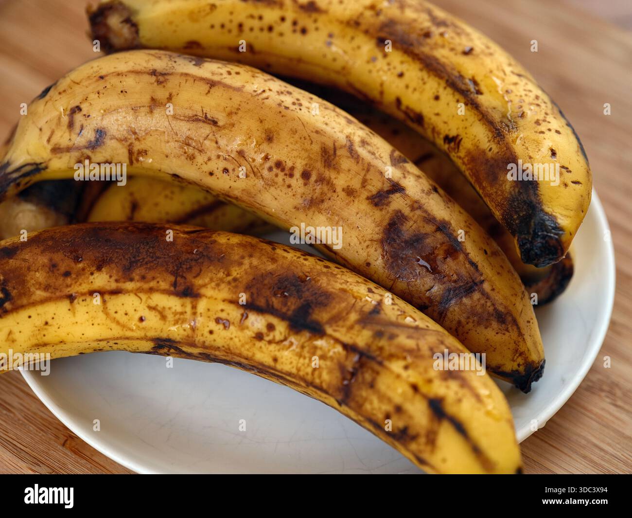 Un mucchio di banane troppo mature con macchie marroni appoggiate su un piatto bianco su un tavolo di legno. Primo piano. Foto Stock
