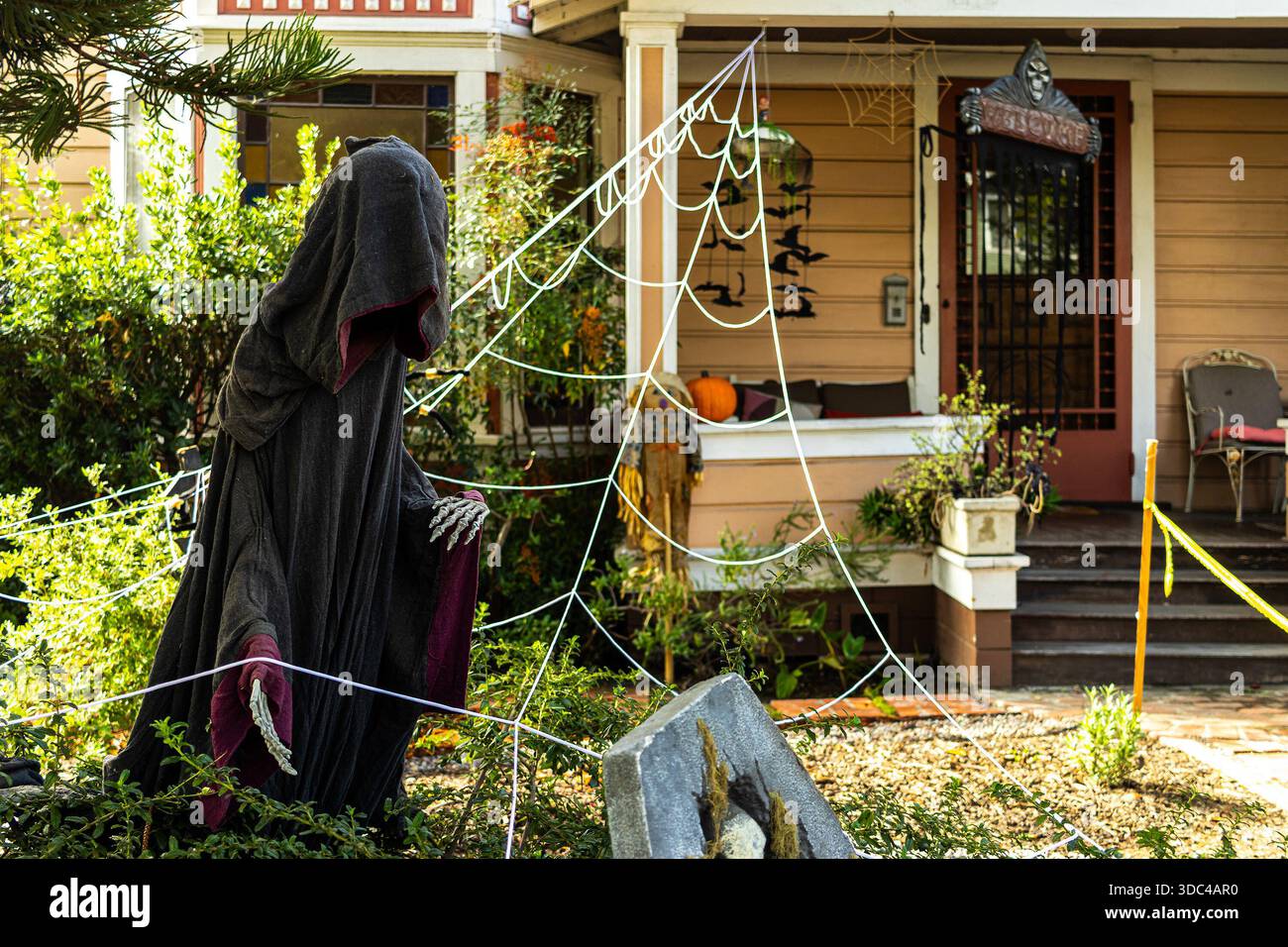Spaventose decorazioni di Halloween in un cortile di periferia con un gesto spettrale con cappuccio e scheletrico verso una lapide e finte ragnatele che coprono il portico Foto Stock